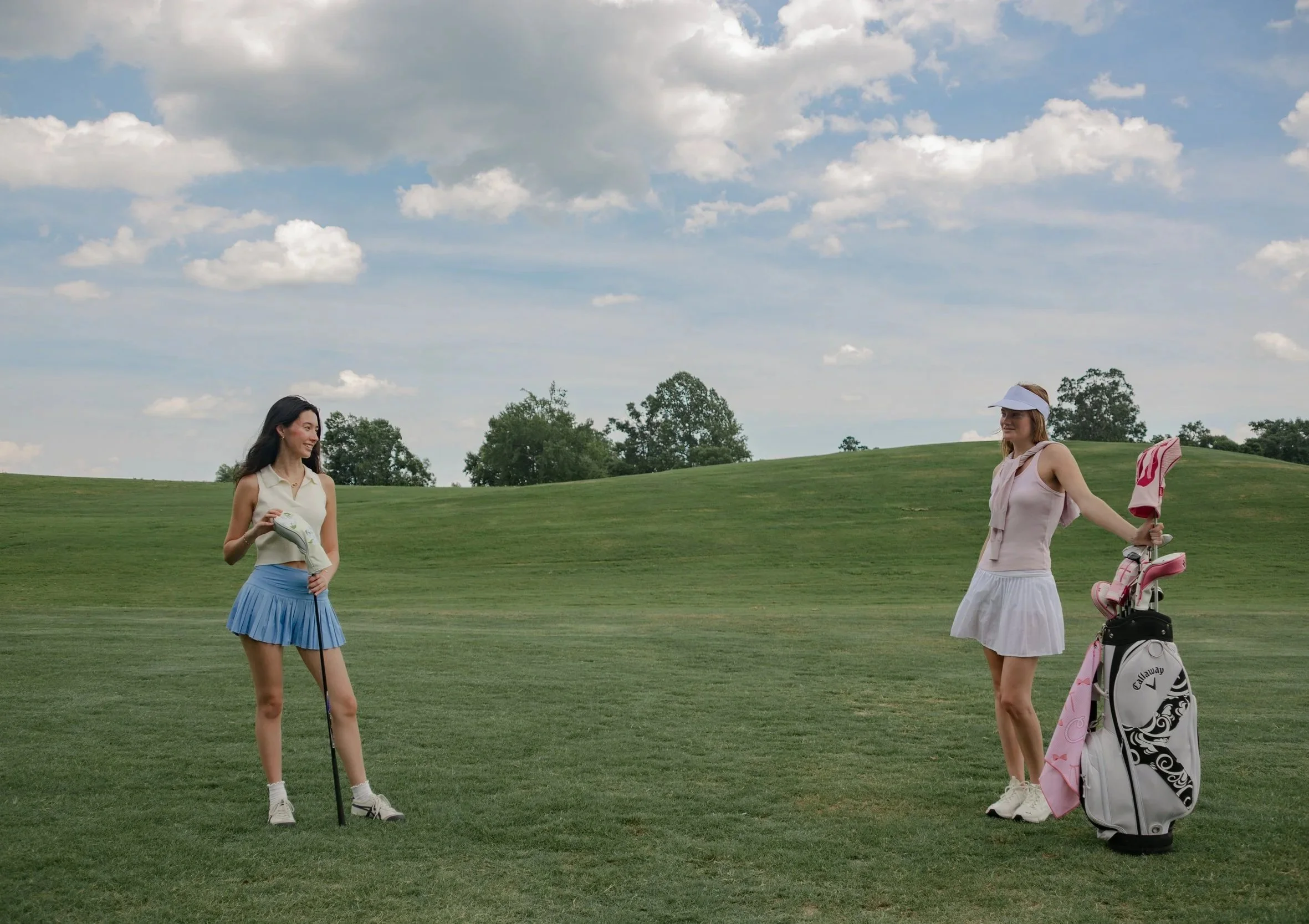 Two women on a golf course, one holding a golf club and the other with a golf bag and cart, under a partly cloudy sky by Rin House Studio in Raleigh, North Carolina.