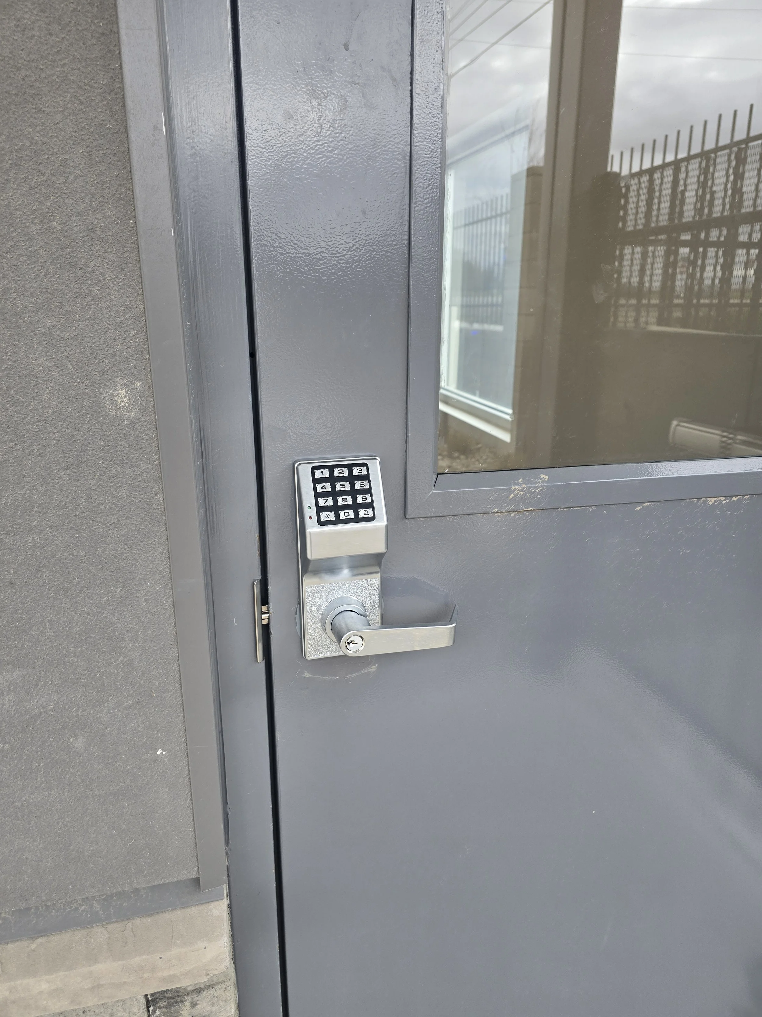 Gray door with a keypad lock and a lever handle, reflected in the glass window above the lock.