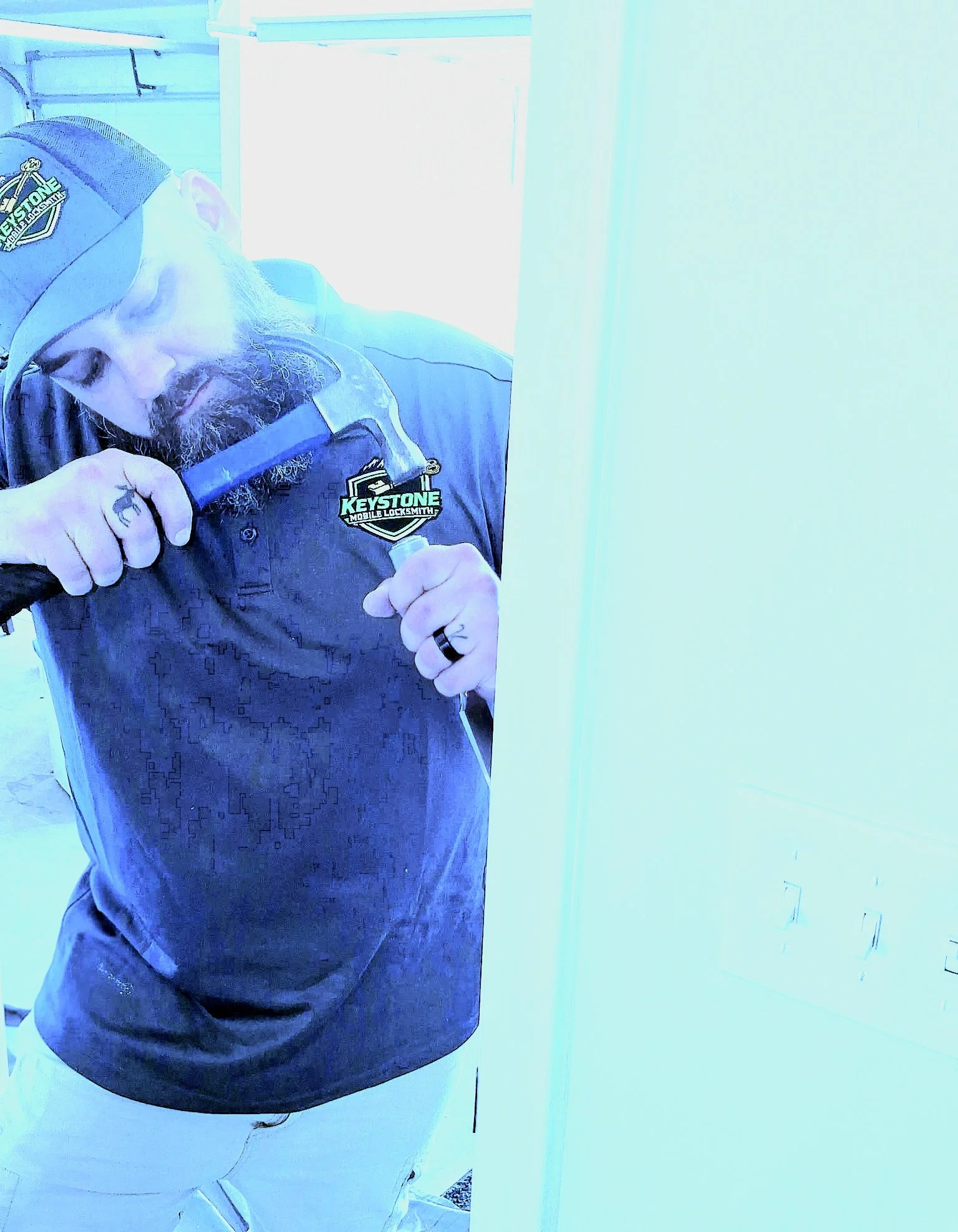 A man with a beard wearing a baseball cap, a dark shirt with a logo that reads 'Keystone Mobile Locksmith,' and white pants. He is looking down and holding a tool in his right hand near his beard.