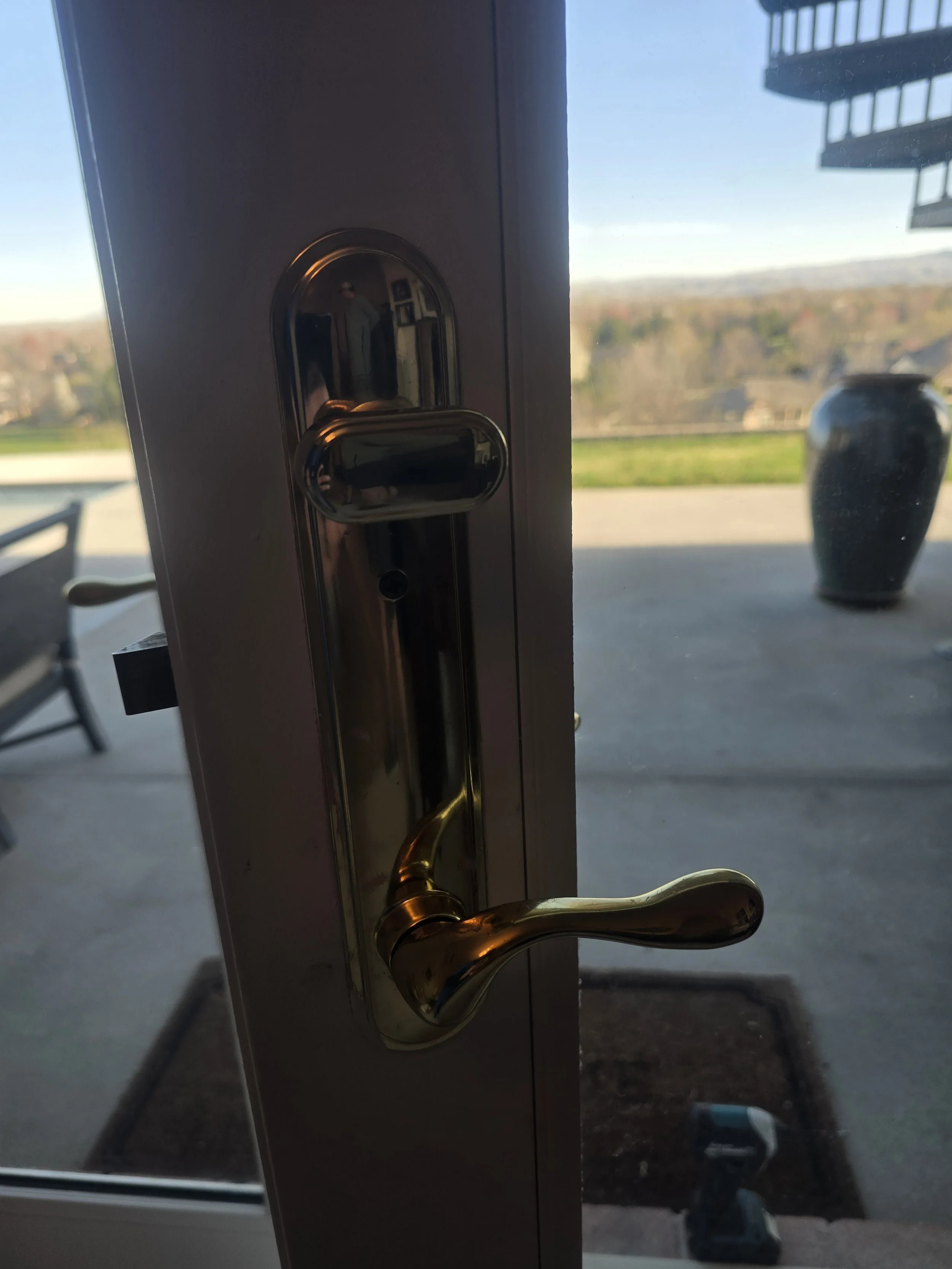 Close-up of a door lock with a golden handle, taken from inside a house or building, with an outdoor view of trees, houses, a parking lot, and a building with balconies in the background.