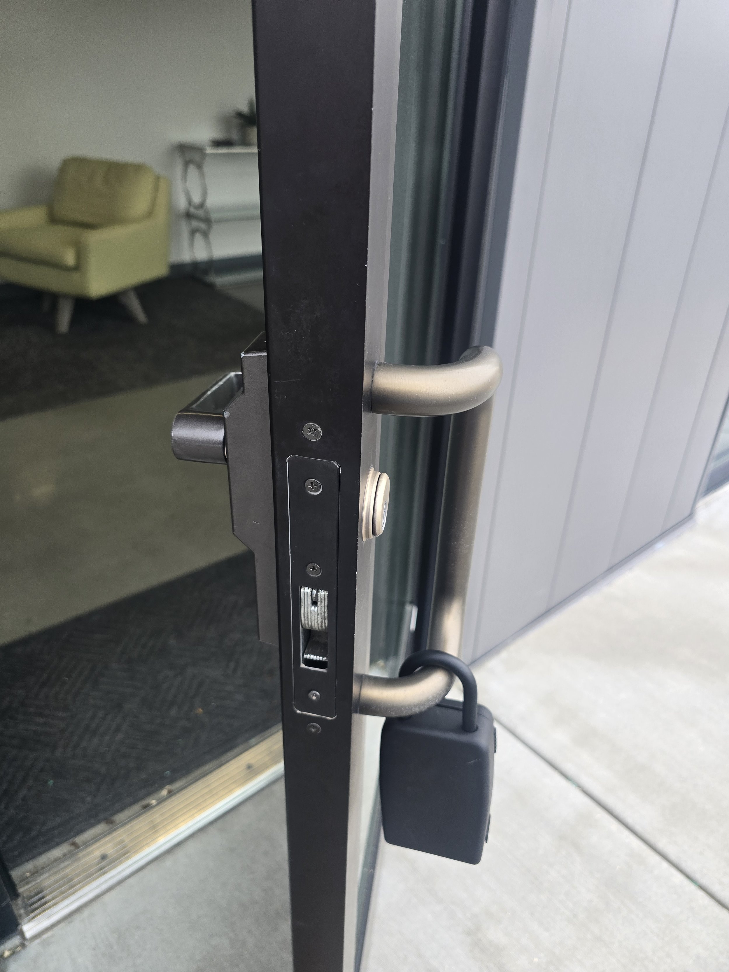Close-up of a door lock and handle on a glass exterior door, with a view into an interior room with a yellow armchair and a white table.