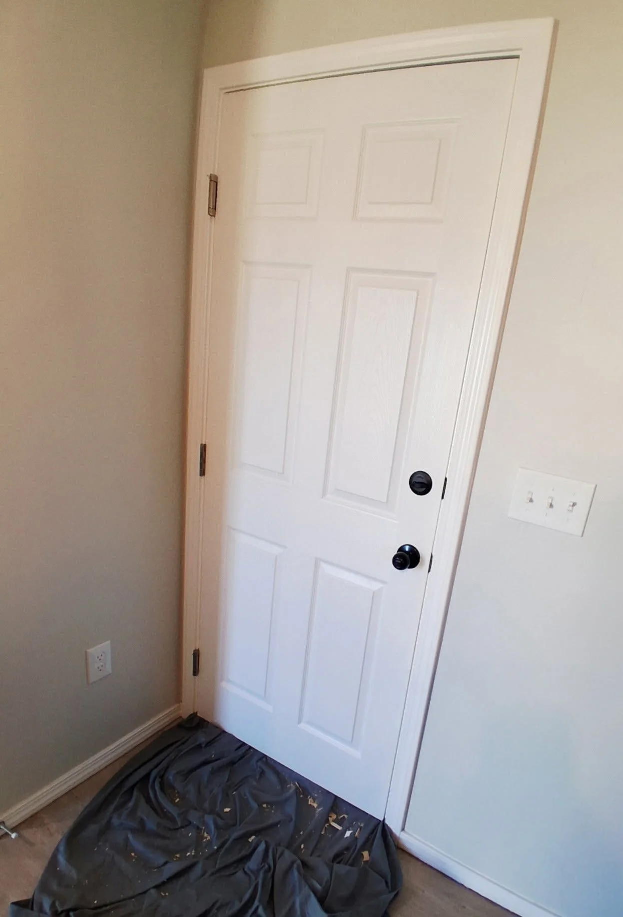 A white interior door with black doorknob and hinges, slightly ajar, in a room with beige walls and a light switch on the right, with a black trash bag and some debris on the floor in front.