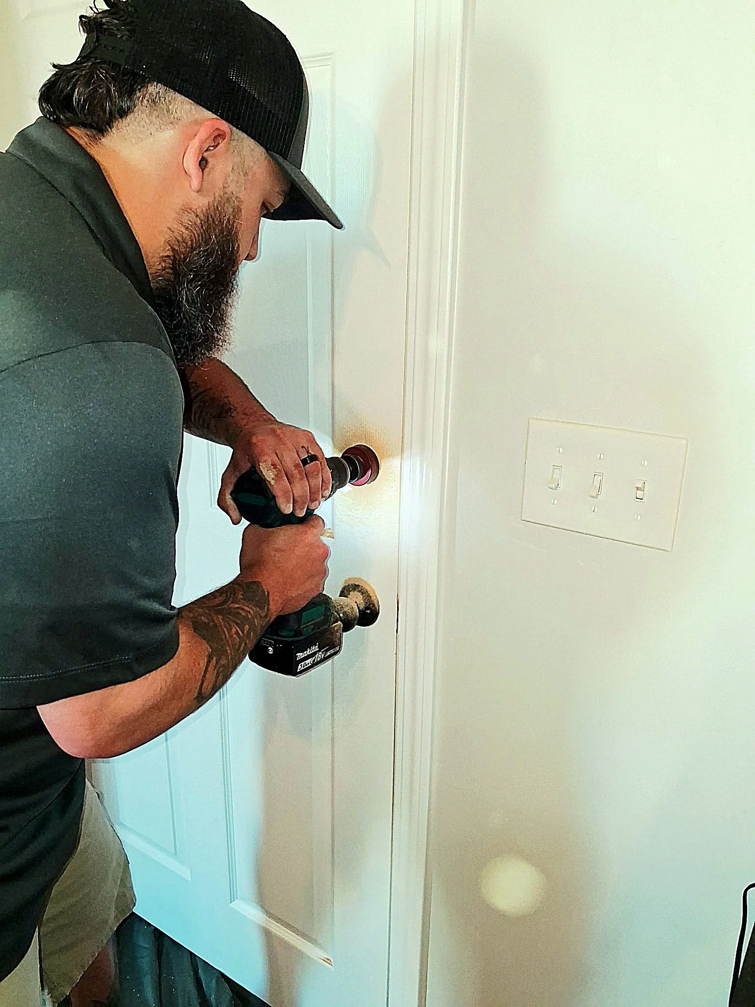Man with a beard and tattoos using a cordless drill on a door in a room with white walls and a light switch.