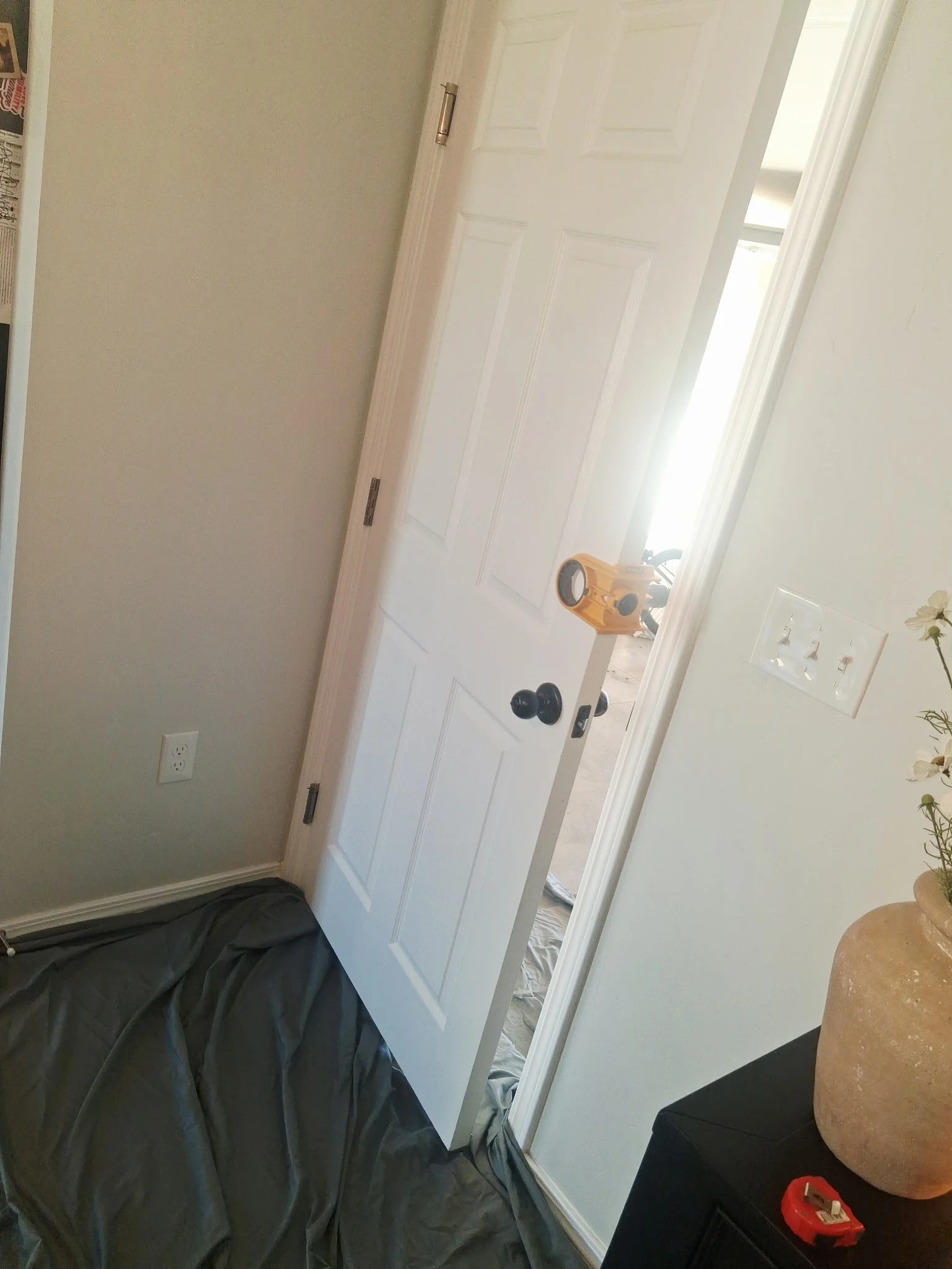 Open white door with black doorknob, revealing a bright room and part of a hallway with wood flooring. A yellow vacuum cleaner is visible through the doorway, and a black bag or object is on the floor nearby.