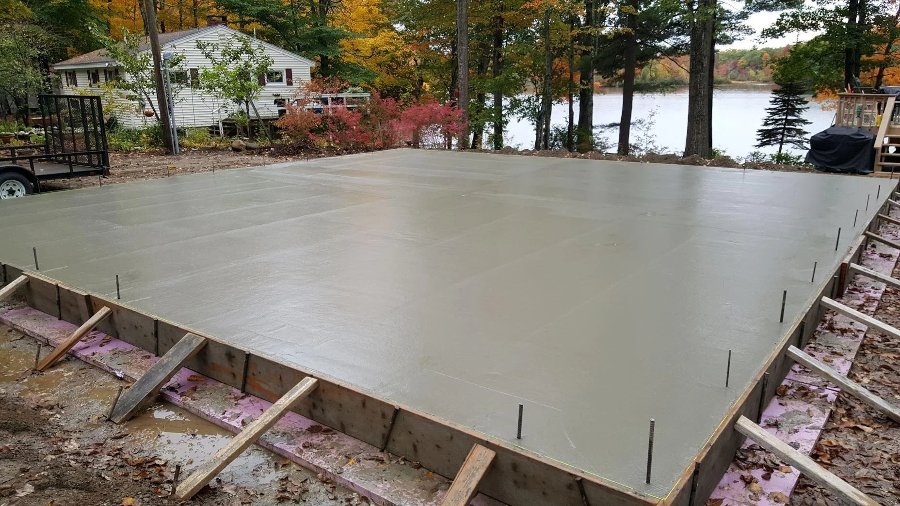 Freshly poured concrete foundation in a backyard with trees and a lake in the background.