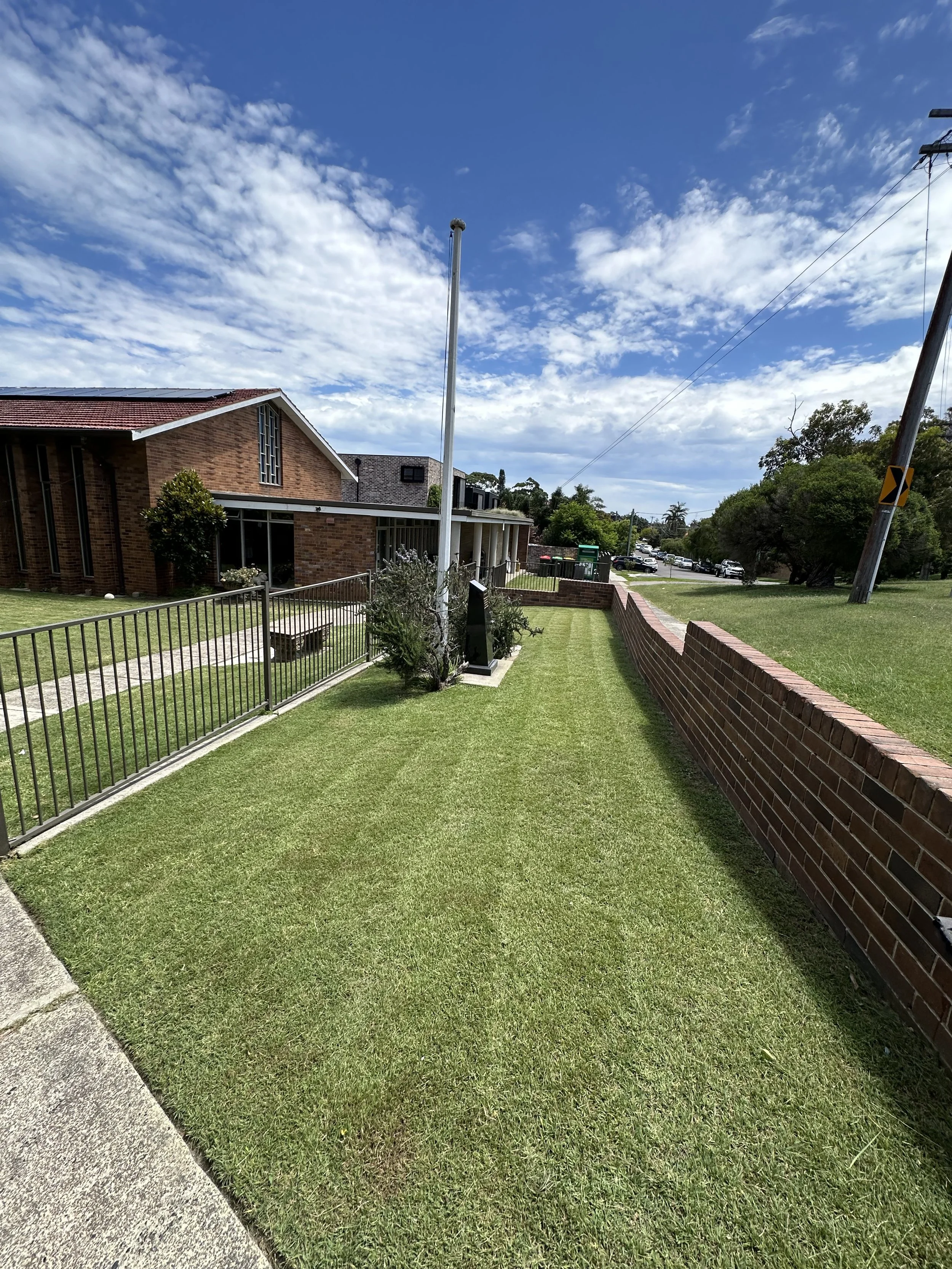A sidewalk and manicured lawn next to a brick wall and metal fence, with a brick building in the background under a partly cloudy sky.