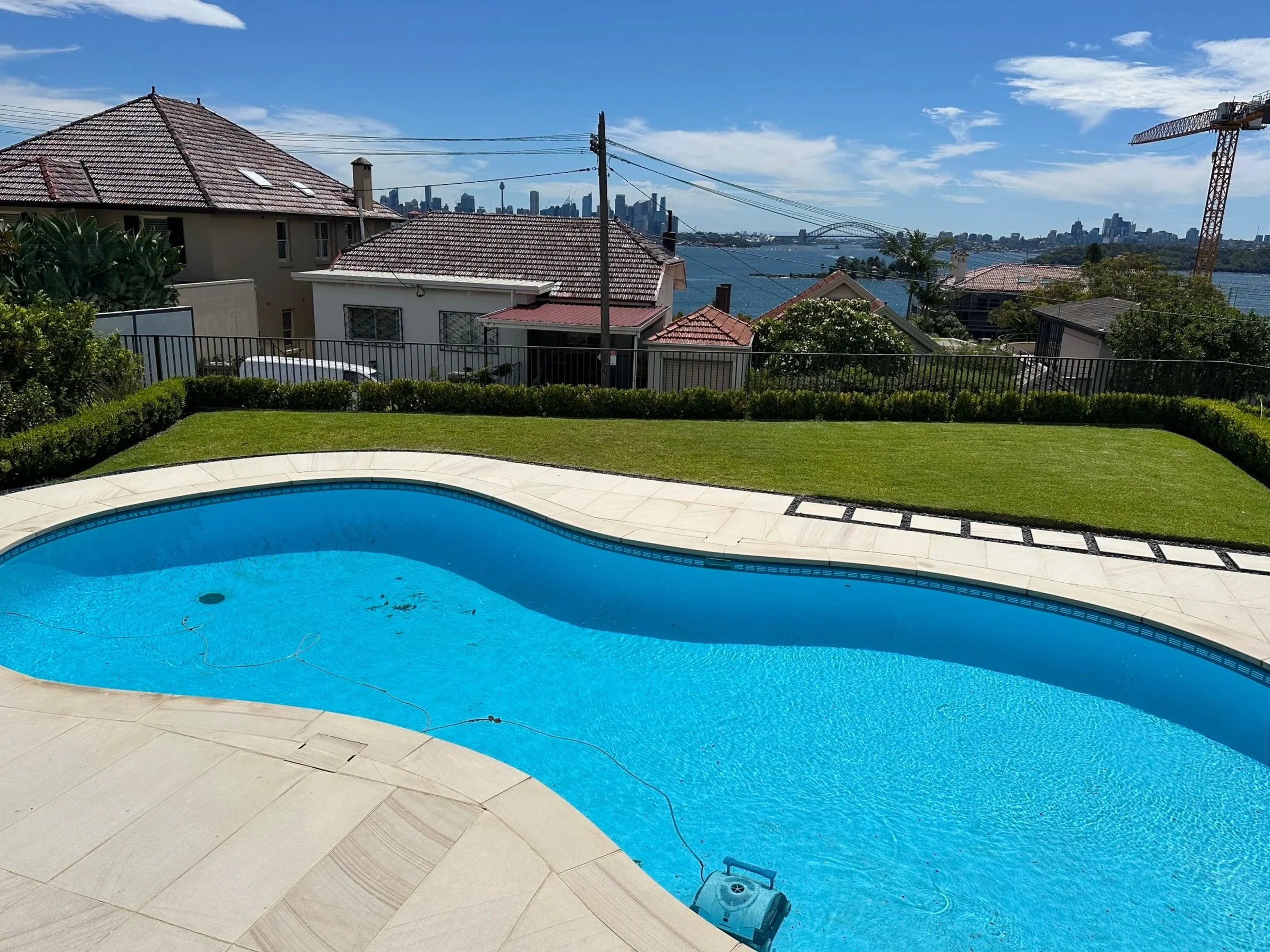Residential backyard with a swimming pool, lush green grass, and a city skyline across a river in the background. House rooftops, power lines, and a construction crane are visible under a partly cloudy sky.