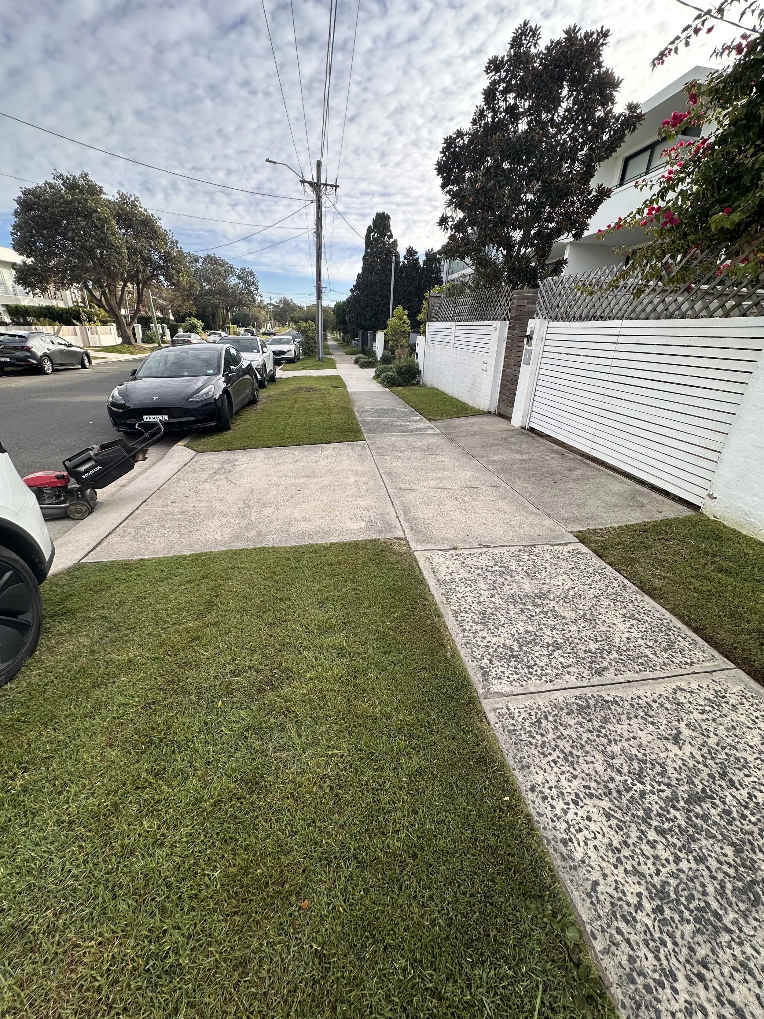 Sidewalk with grass patches, parked cars along the street, trees, residential buildings, and a partly cloudy sky.