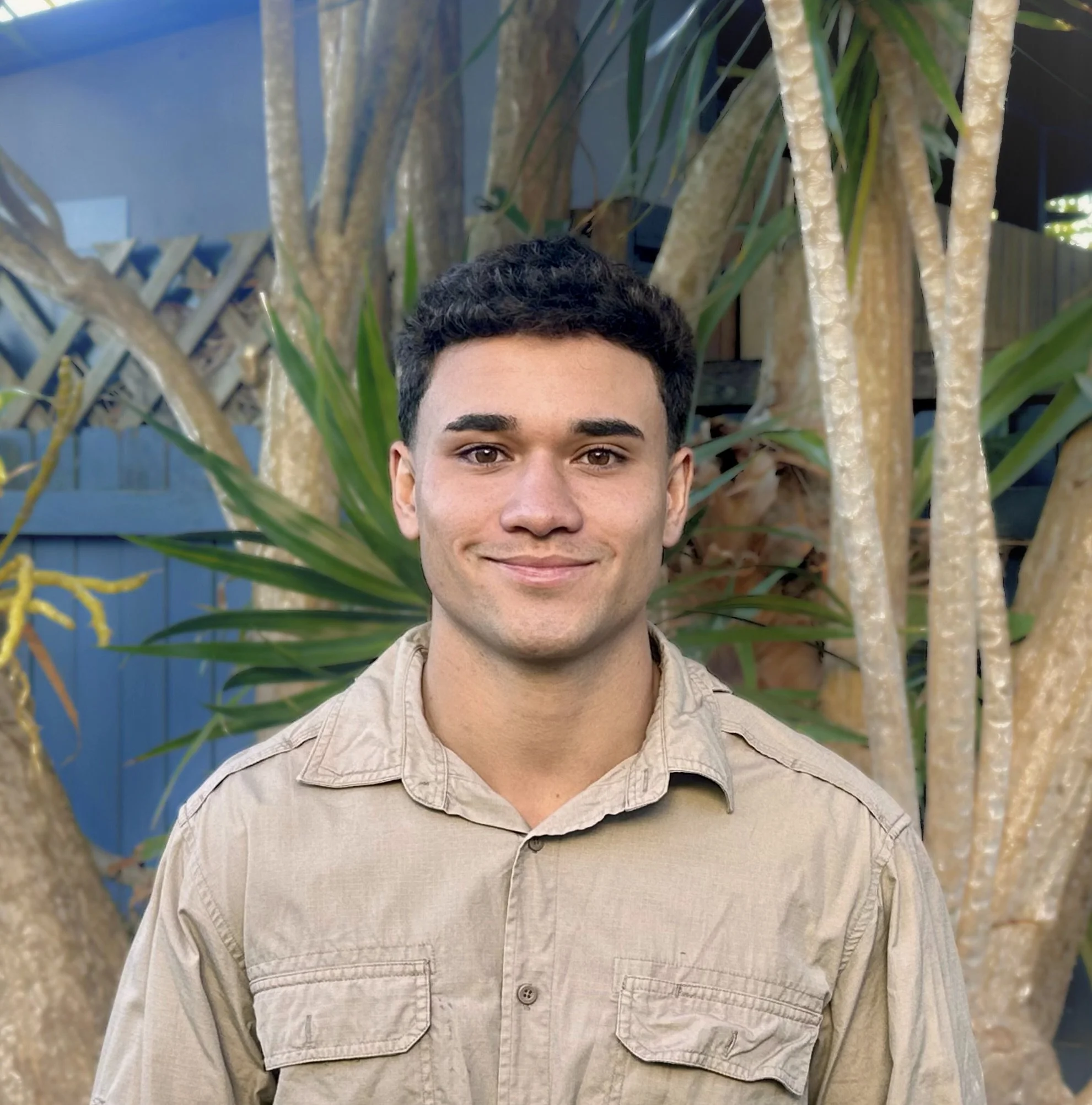A young man with short dark hair and a slight smile, standing outdoors in front of a large plant with thick branches and green leaves.