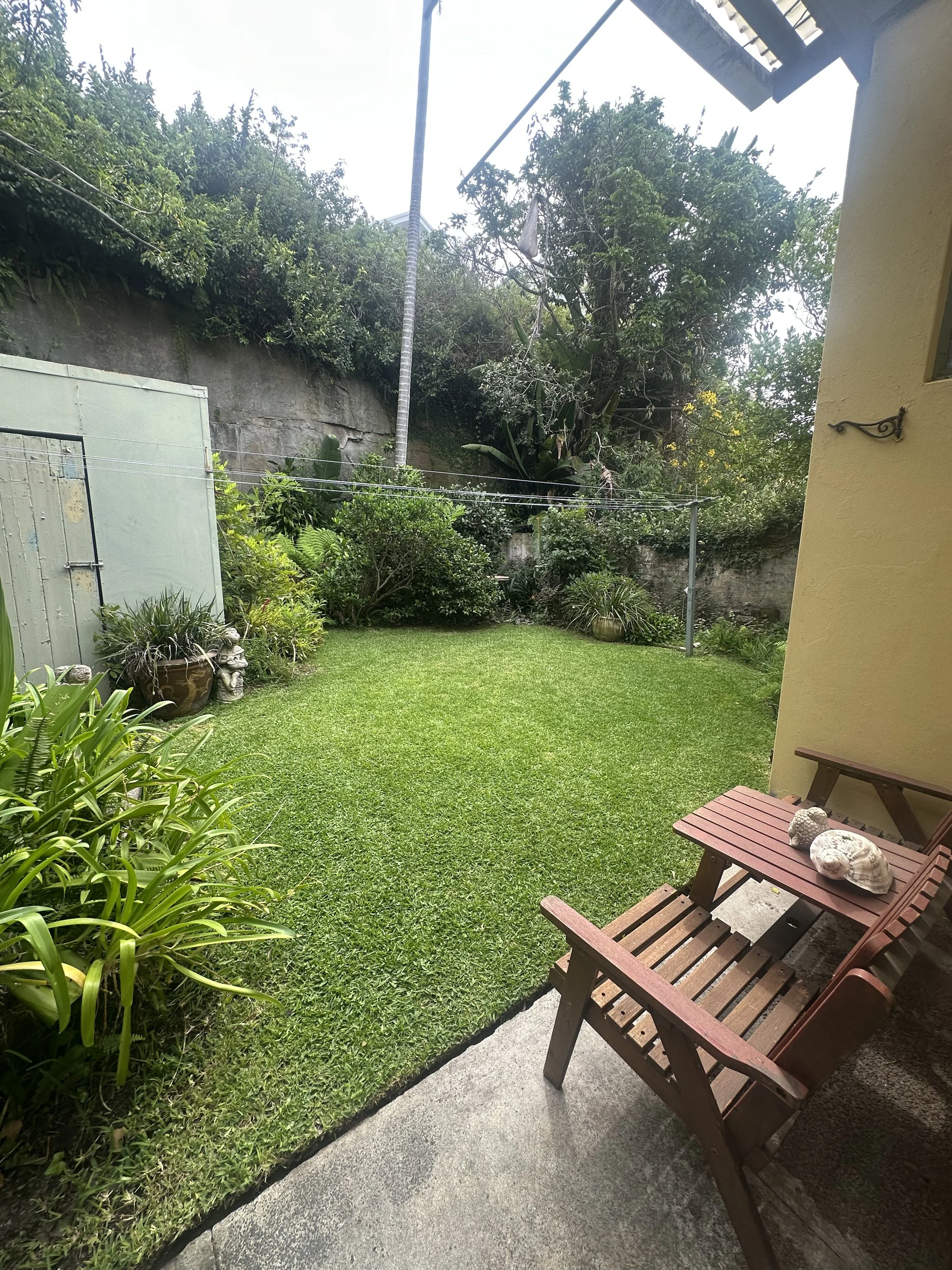 View of a lush backyard with green grass, plants, and trees, a clothesline with hanging laundry, and a wooden bench with seashell decorations on a patio.