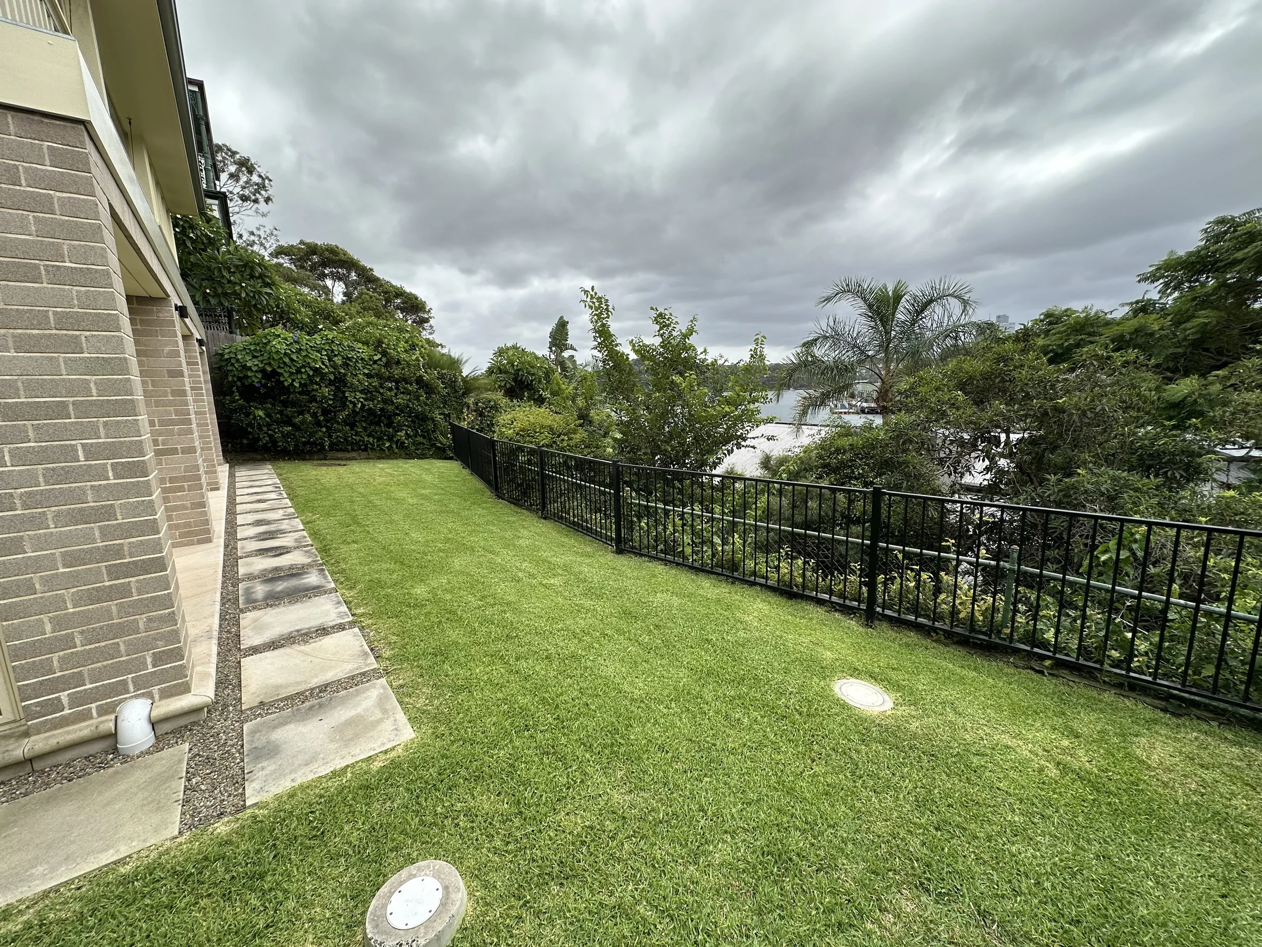 View of a backyard with green grass, various trees, bushes, and a black fence, with cloudy sky overhead, part of a house wall with brick and siding visible on the left.
