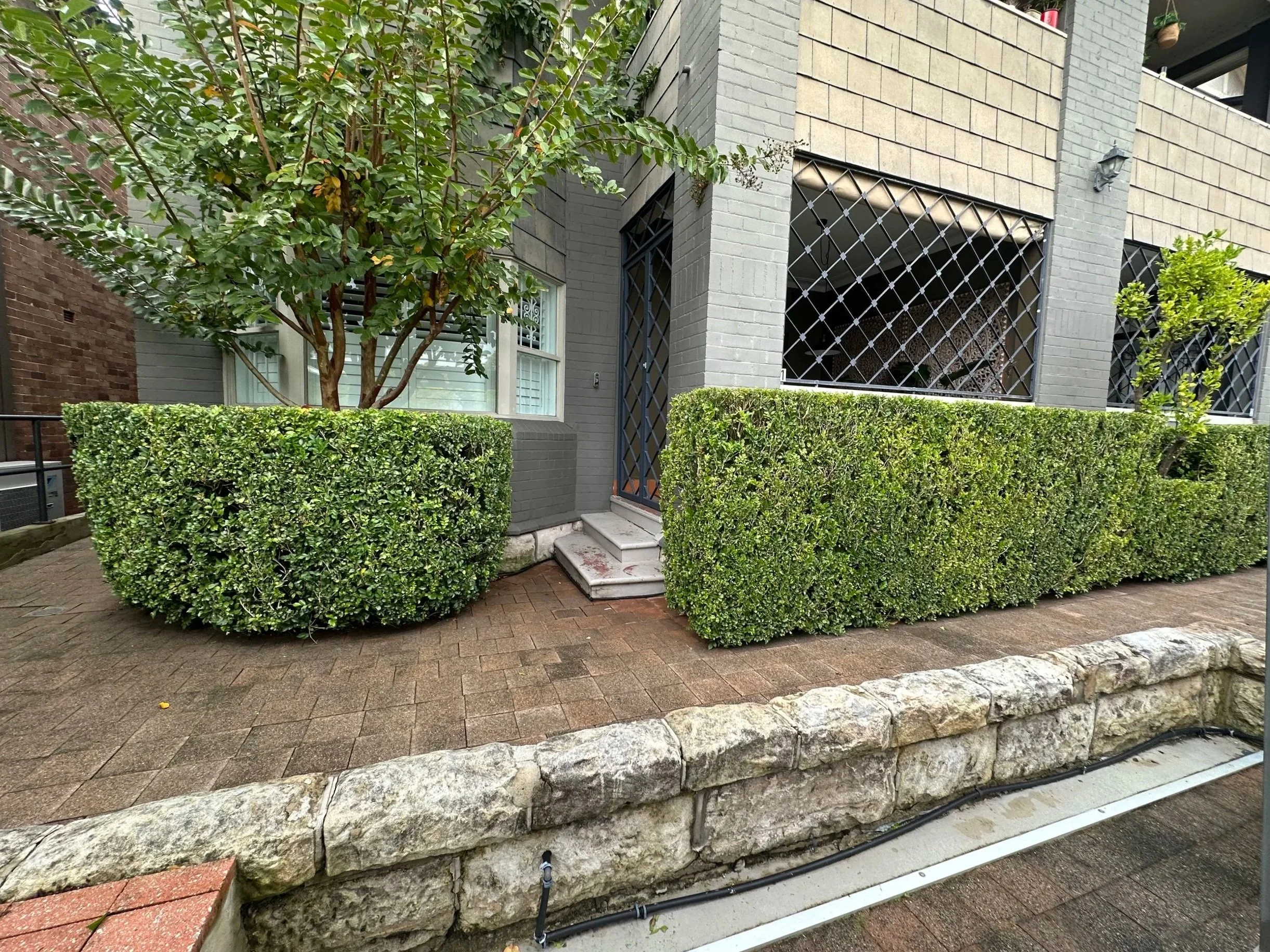 A modern multi-story building with a gray and beige brick facade, surrounded by well-maintained green bushes and trees, with steps leading to an entrance.