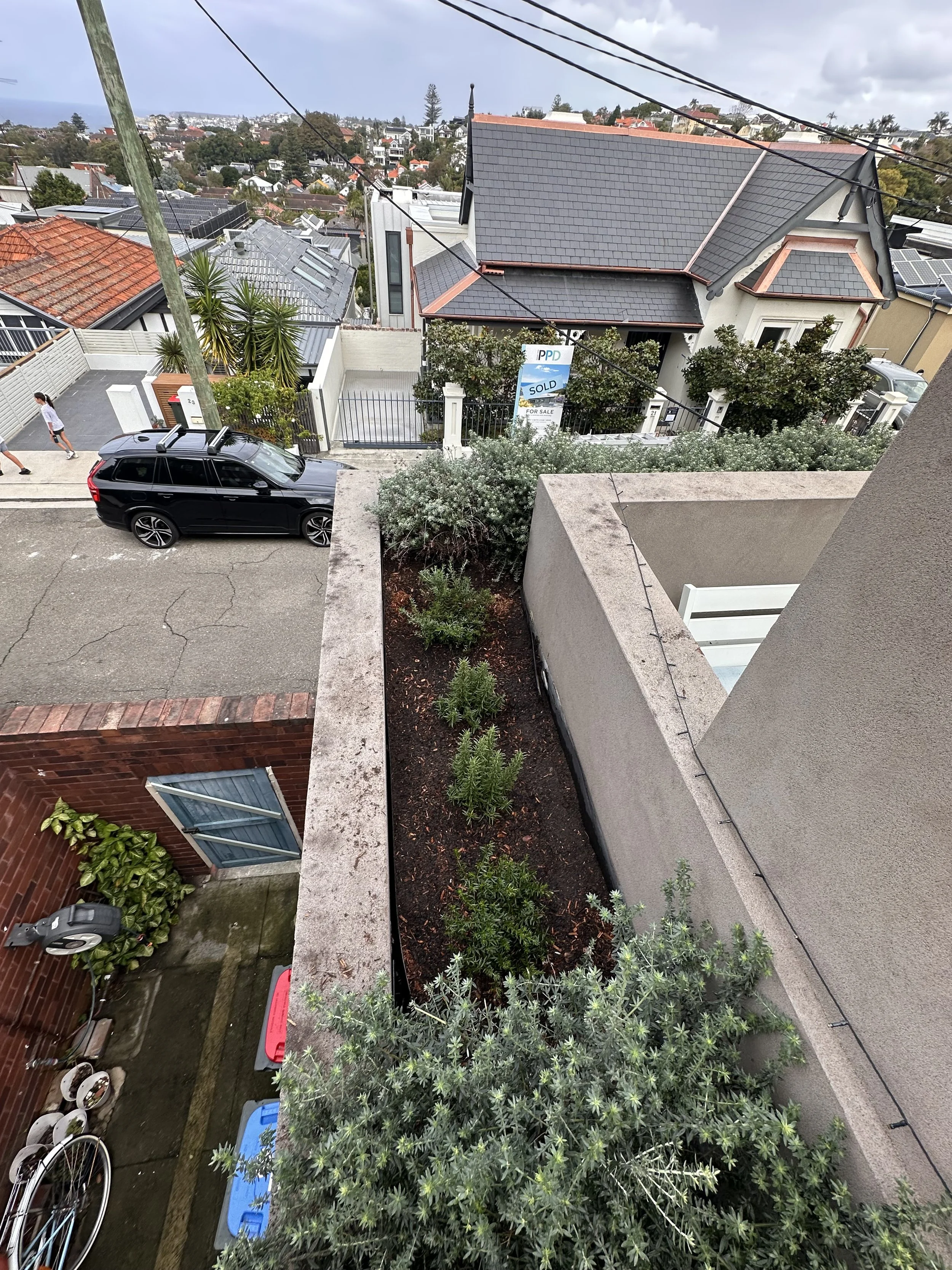 View from a balcony showing a residential neighborhood with houses, a street, a black car, and greenery under a cloudy sky.