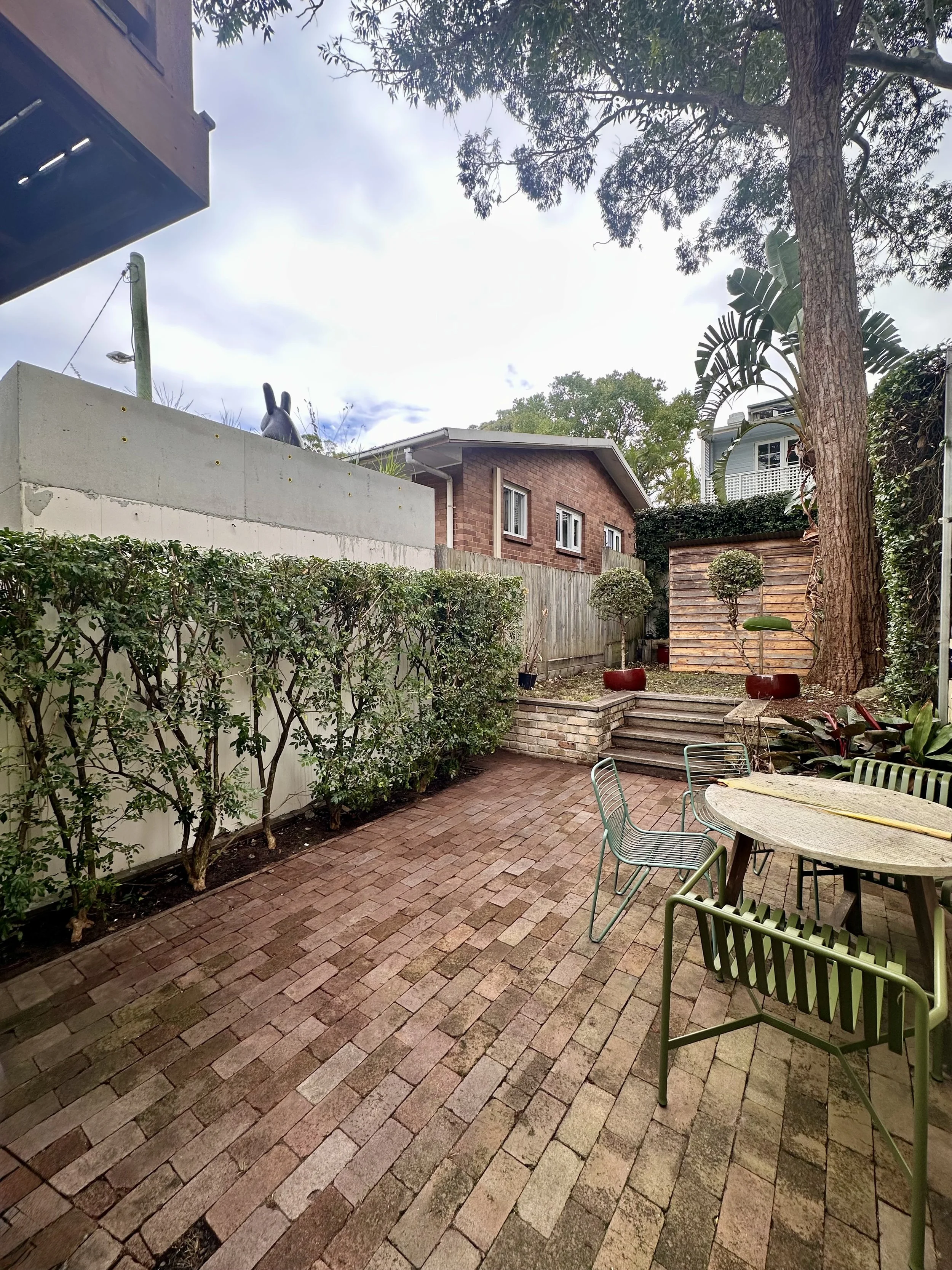 A backyard patio with brick paving, a round table with green chairs, potted bushes, a large tree, a small shed, and a brick house in the background.