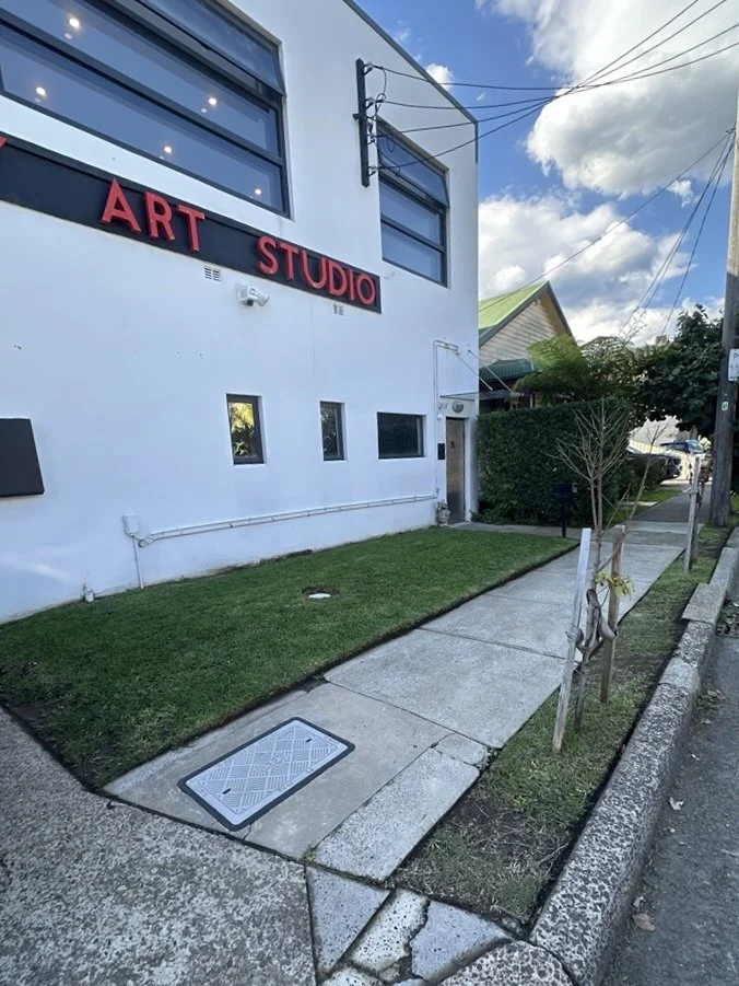 Exterior of an art studio building with a white facade, large windows, and red sign reading 'ART STUDIO'. There is a sidewalk, small grass patch, young trees, and a clear blue sky with some clouds.