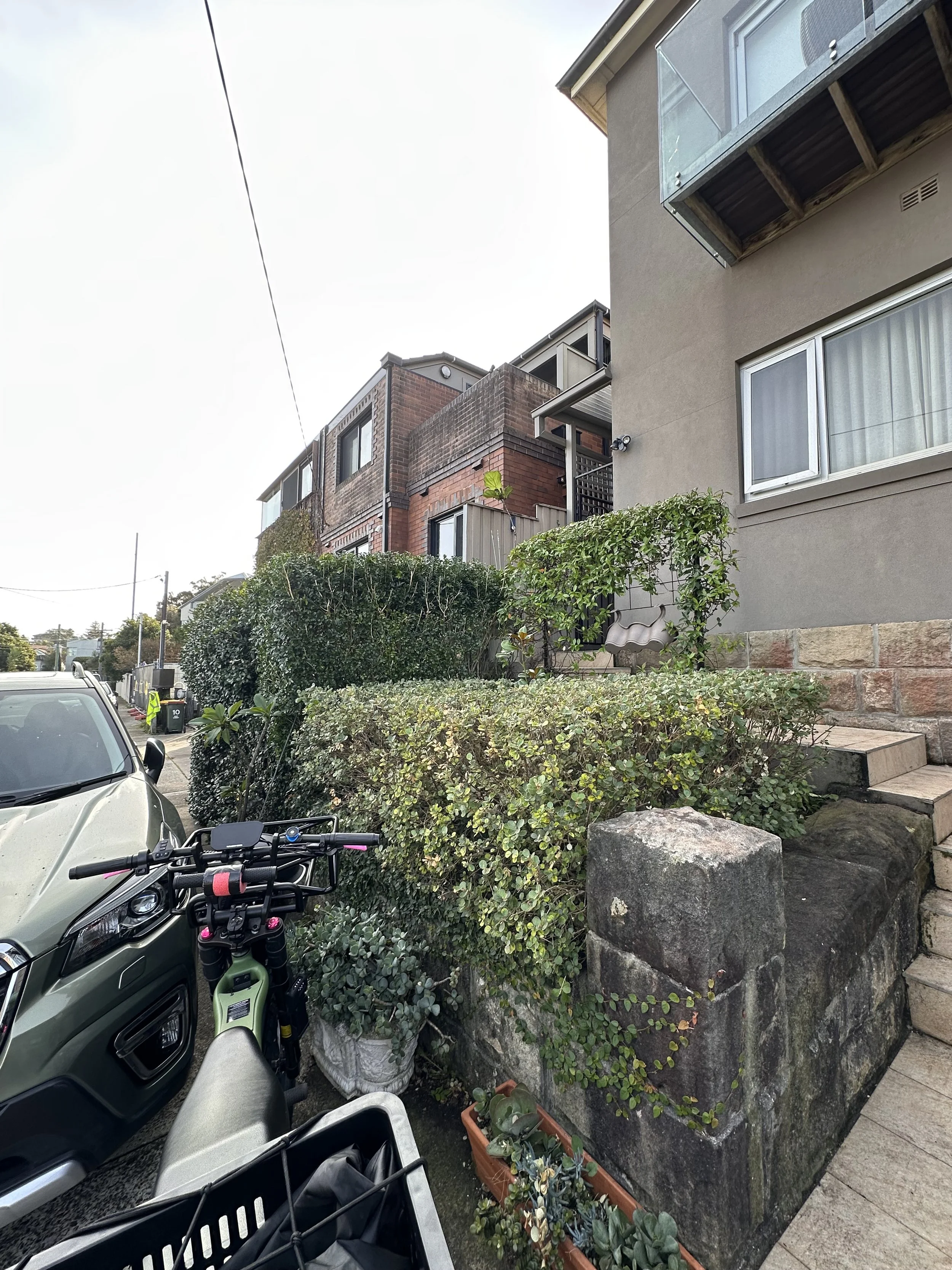 Residential sidewalk with planters, bushes, parked car, and bicycle in front of houses with windows and balconies.