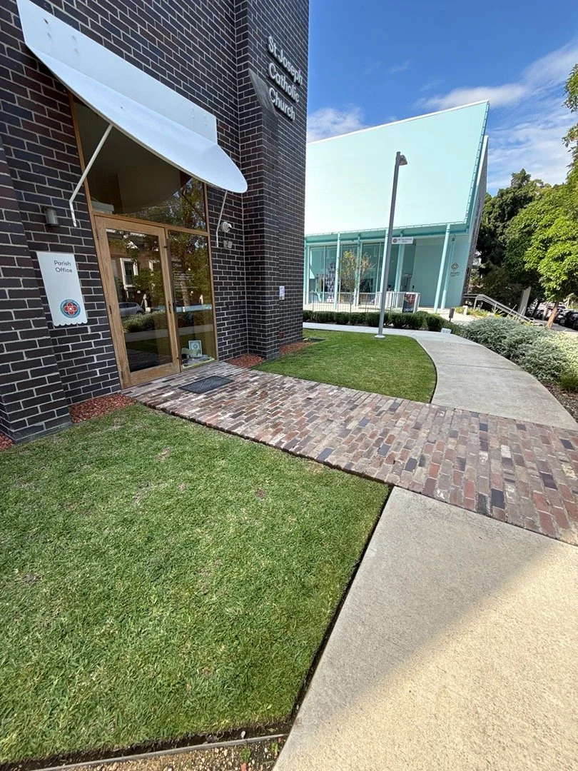 Exterior view of St. Joseph Catholic Church with brick and modern architecture, including a brick walkway, grass, and a contemporary entrance with a white awning and glass door.