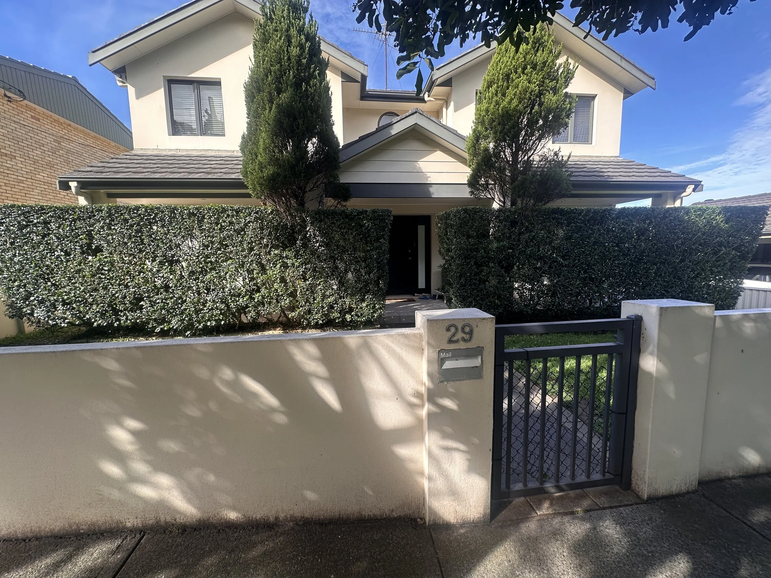 A two-story house with a beige exterior, gray roof, and large windows, partially obscured by tall, dark green trees and a neatly trimmed hedge, with a black metal gate, the house number 29, and a mailbox in the front yard.