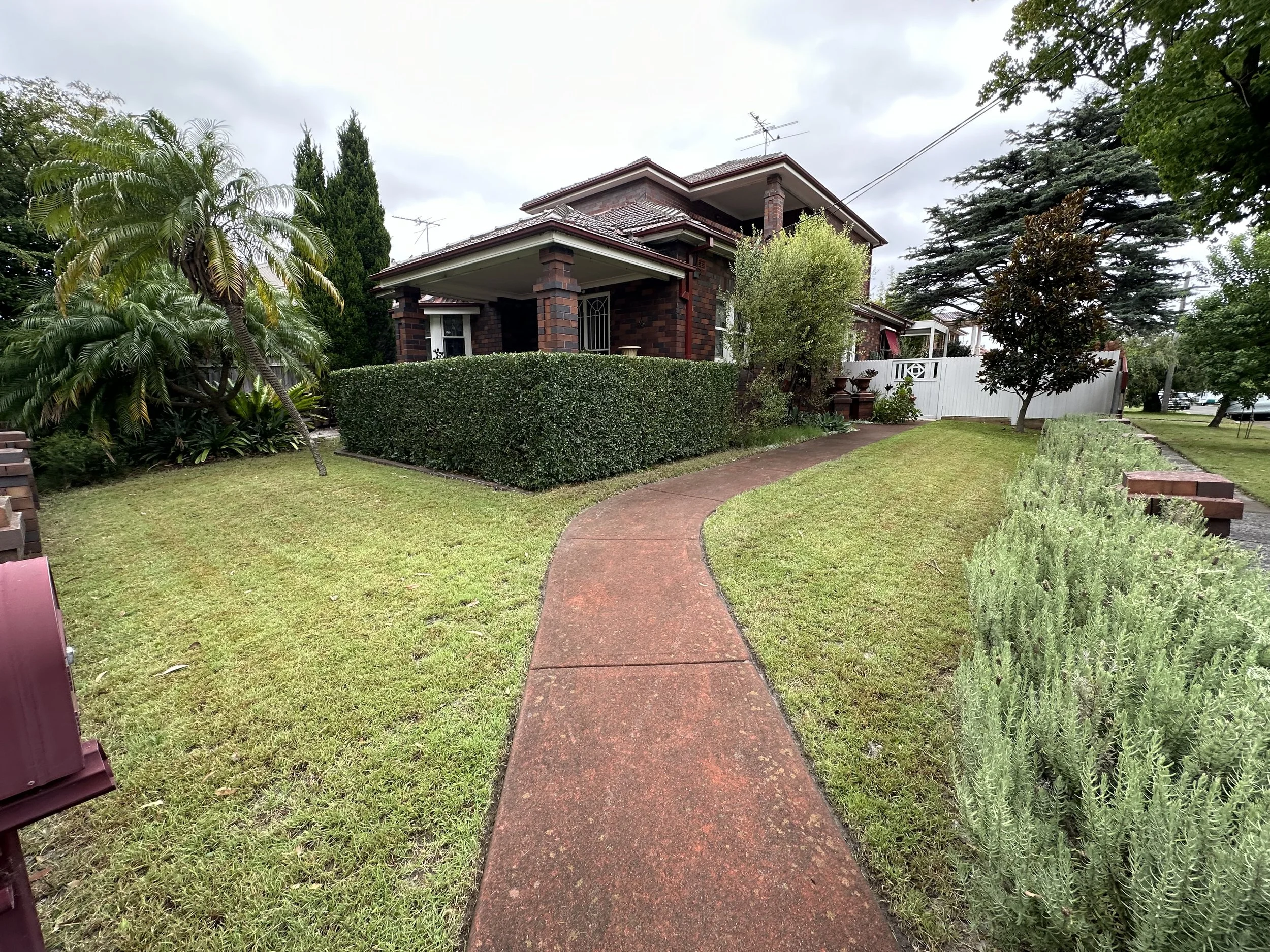 A brick house with a covered front porch, surrounded by lush greenery including a palm tree, bushes, and large trees. A red walkway leads to the front of the house. The sky is mostly cloudy.