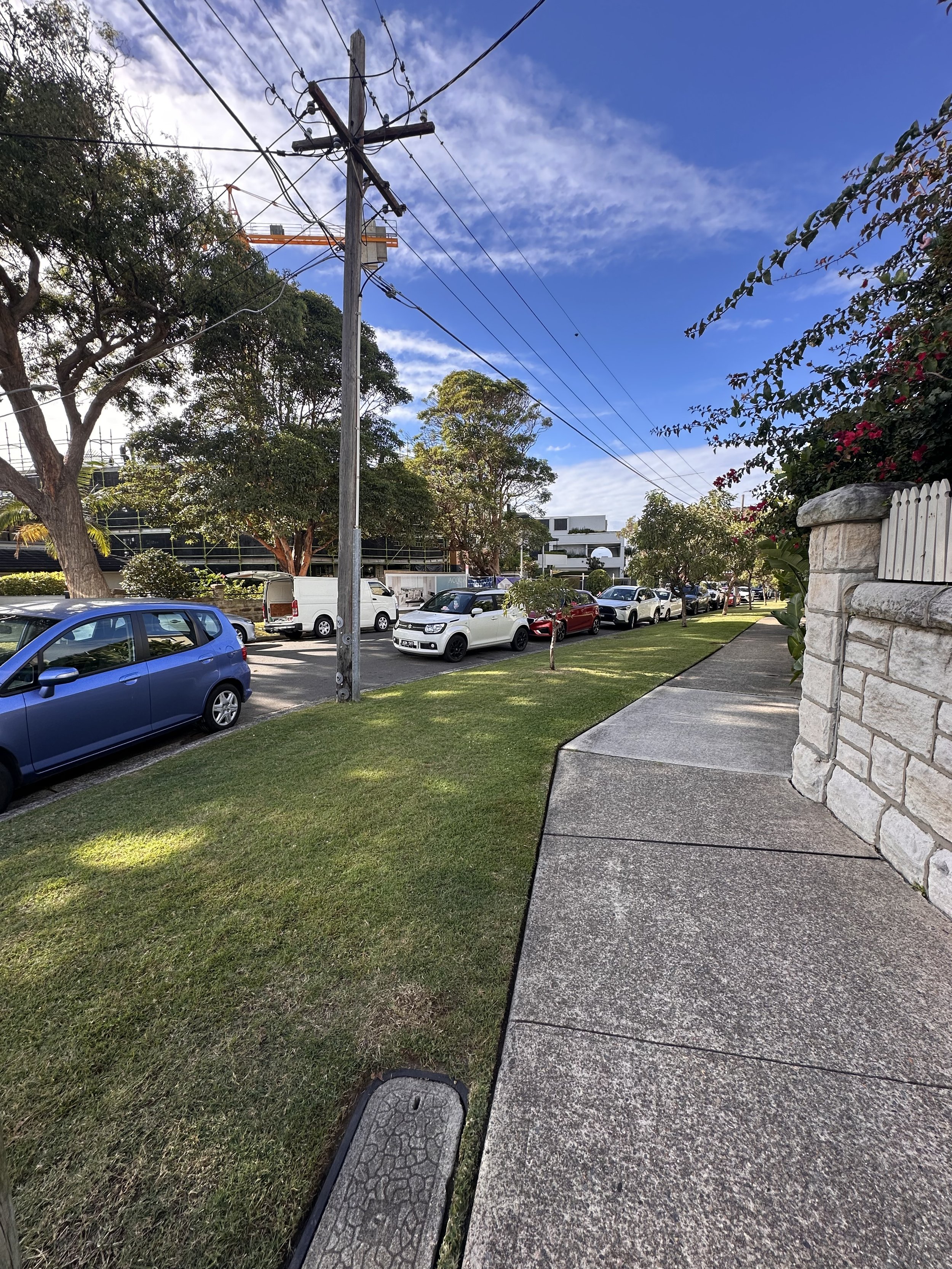 A residential street scene with a sidewalk on the right, a grassy lawn, parked cars, trees, power lines, and a blue sky with scattered clouds.