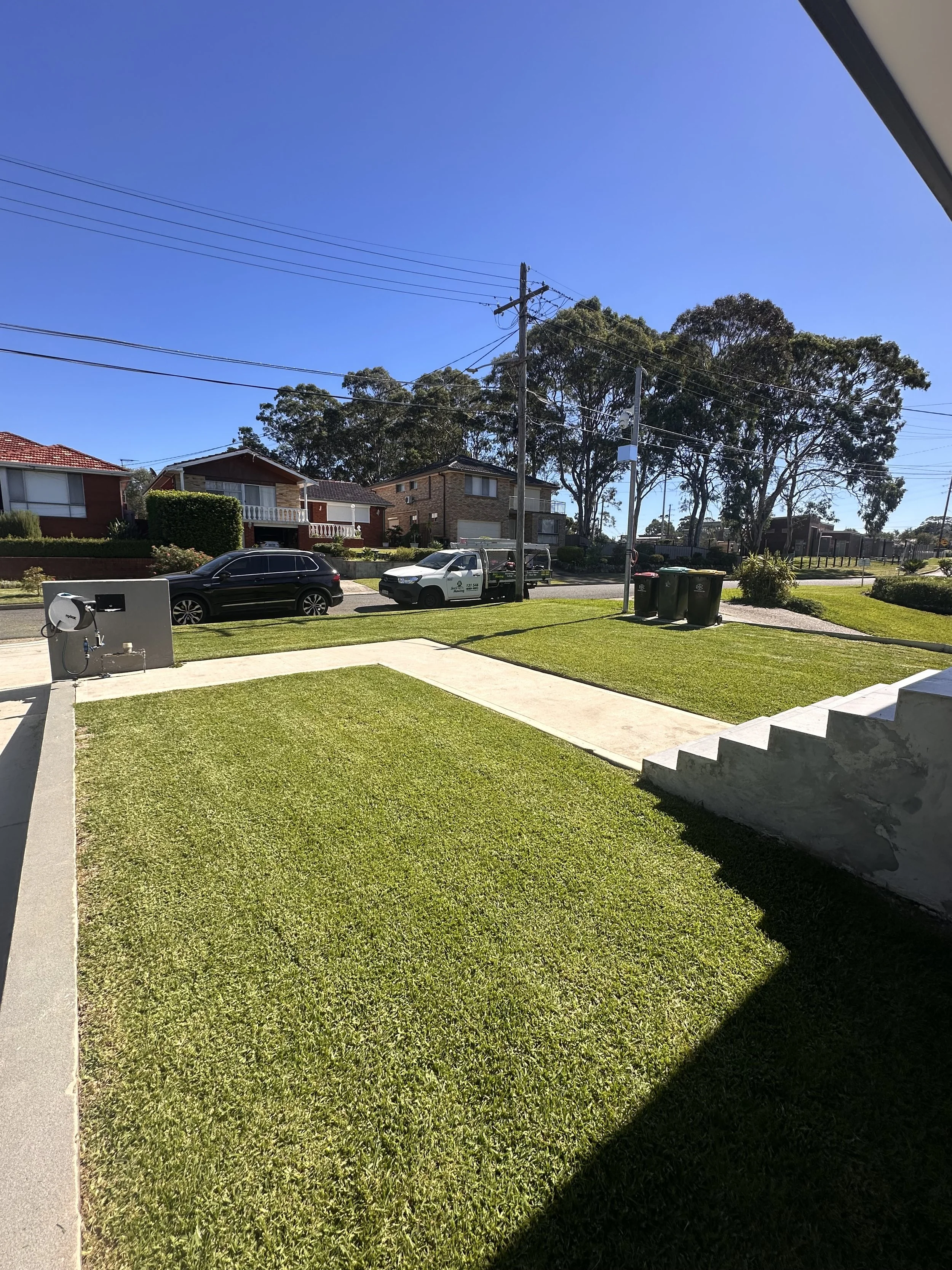 A suburban neighborhood with houses, trees, a parked car, a utility pole, and trash bins on a sunny day with a clear blue sky.