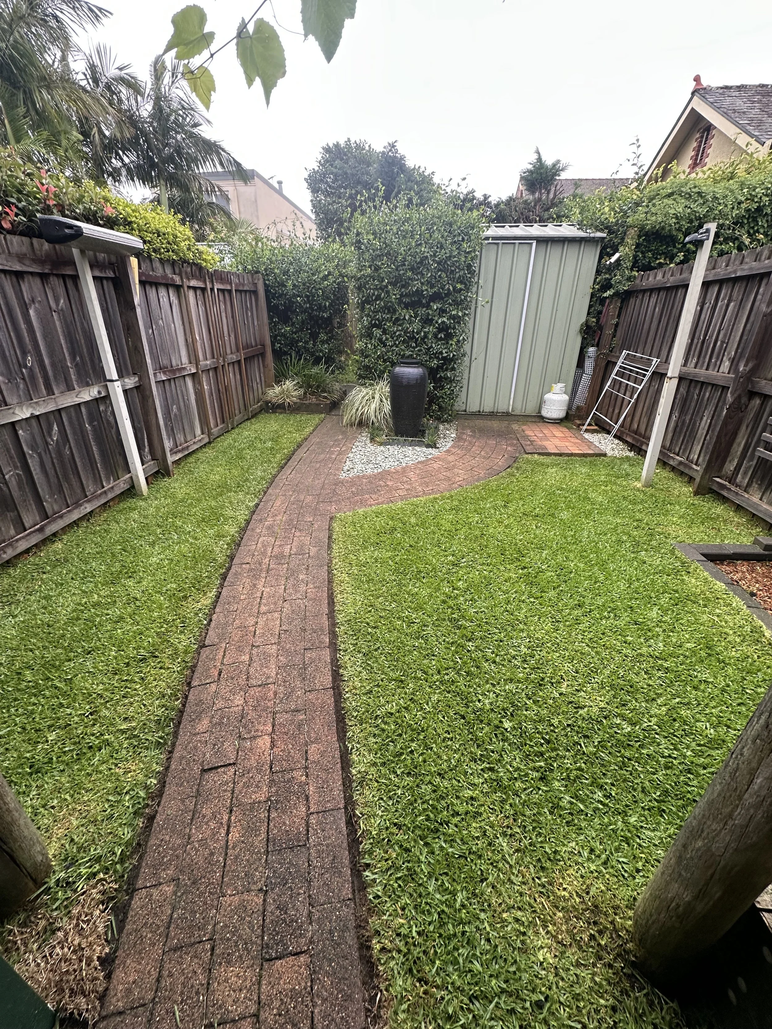 A small backyard with a brick pathway, green grass, a garden shed, and various plants and shrubs, enclosed by wooden fences.
