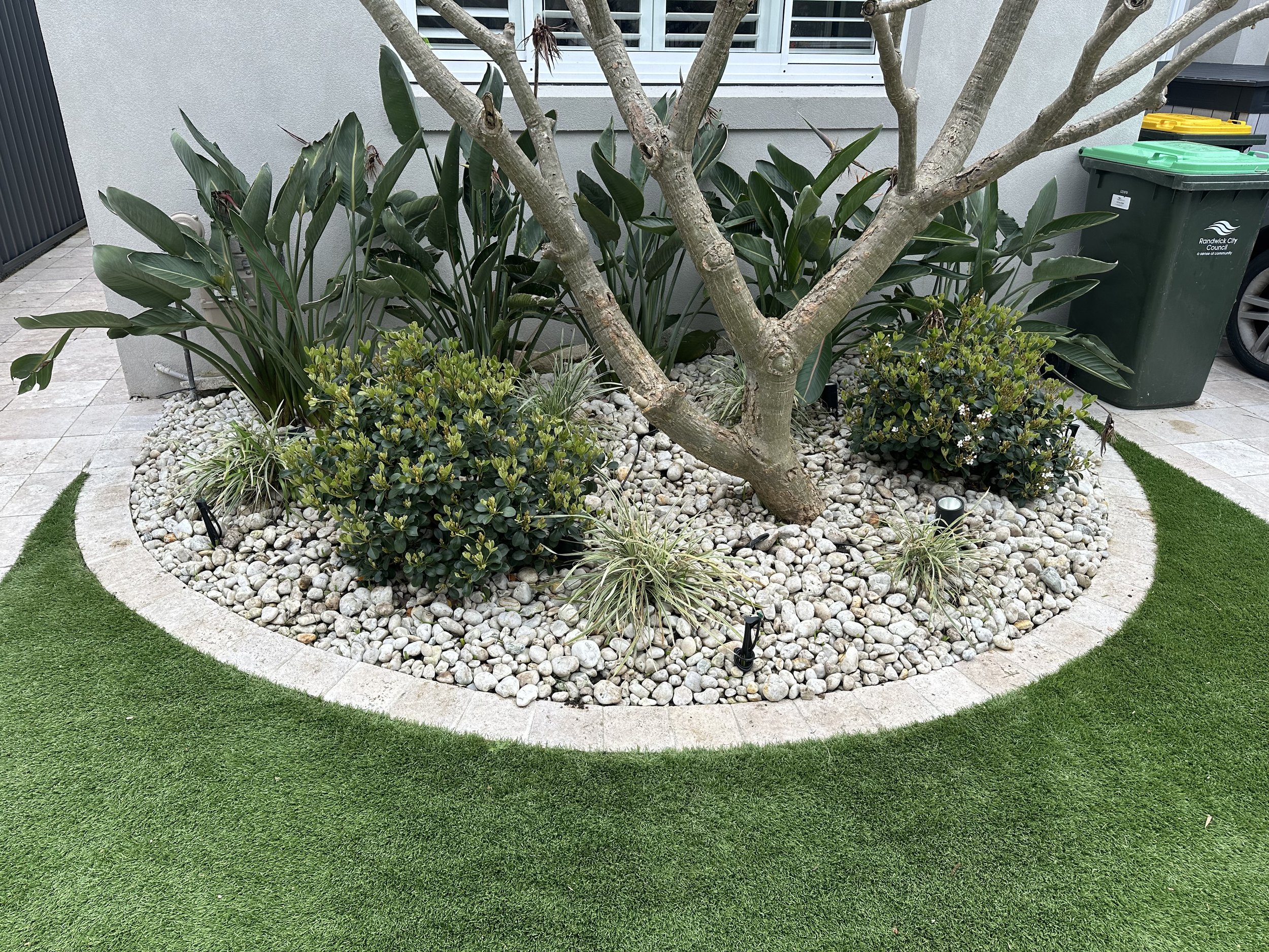 A landscaped yard with a small tree surrounded by white rocks, green plants, and artificial turf edging, with a gray wall and trash bins in the background.