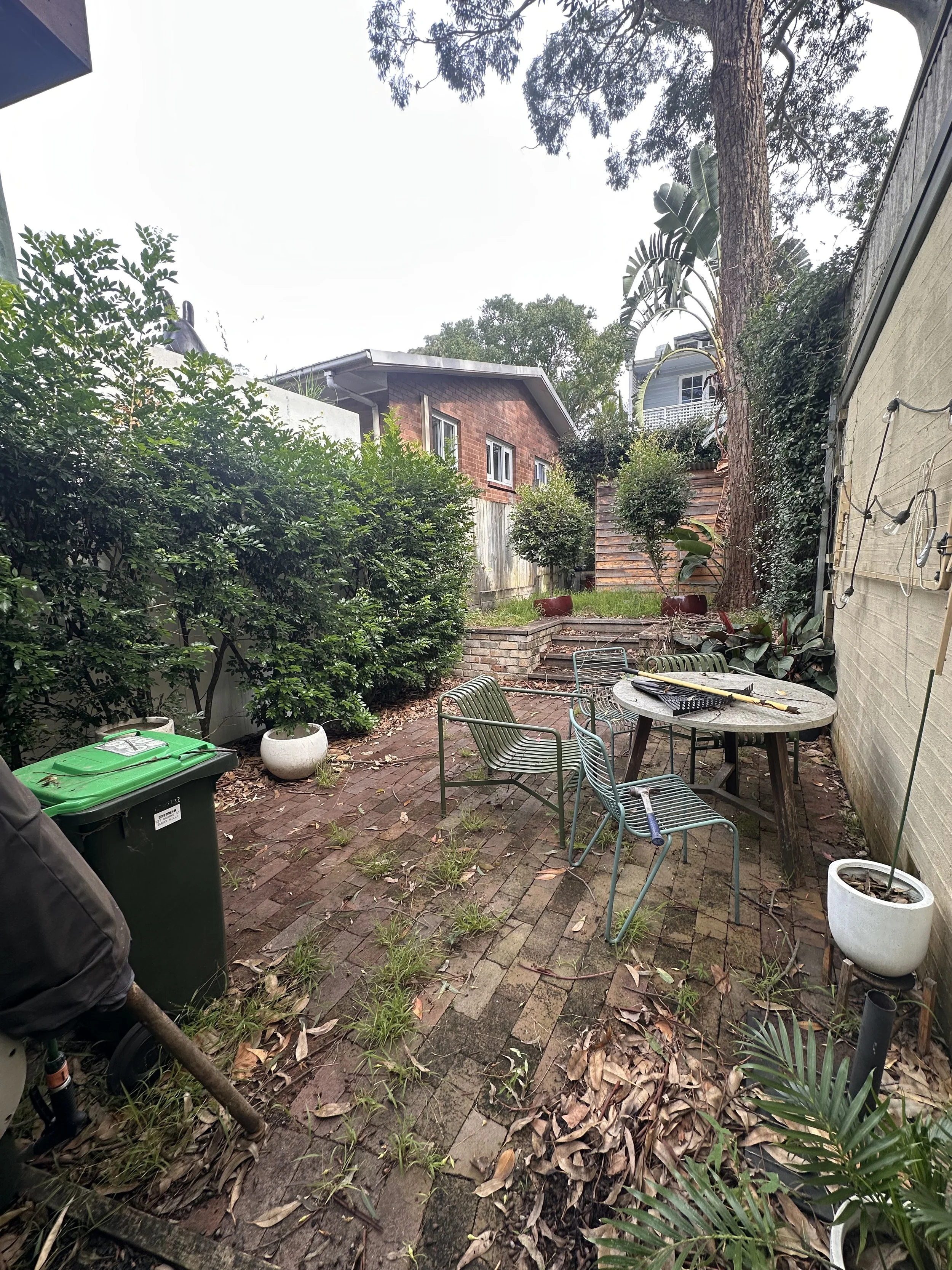 A backyard patio with a dining table, four chairs, and scattered leaves on the brick ground. There are various plants and trees along the edges, and a two-story brick house with a wooden fence in the background. Garden tools and a green trash bin are also visible.