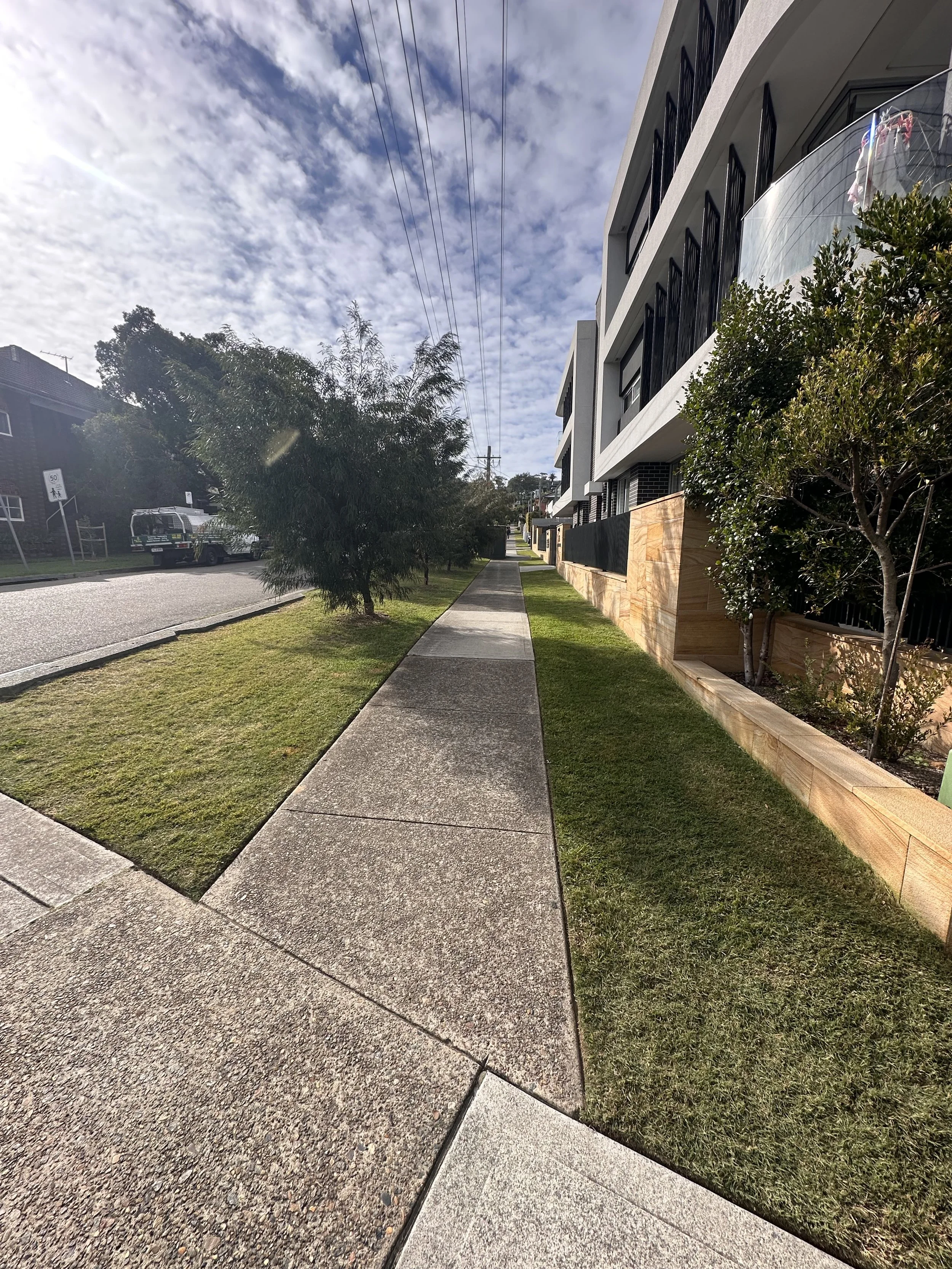 A wide, straight sidewalk along modern residential apartments with glass balconies and a wooden fence, flanked by green grass and trees, under a partly cloudy sky with power lines overhead.