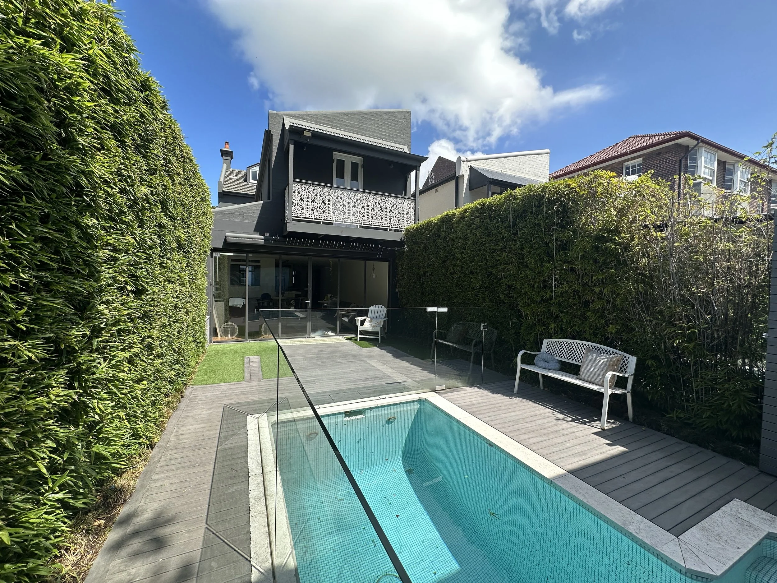 Backyard with a small rectangular swimming pool gradient blue in a wooden deck area, surrounded by a green hedge on the sides and outdoor furniture including a white bench and chairs. There are multistory residential buildings in the background under a partly cloudy sky.