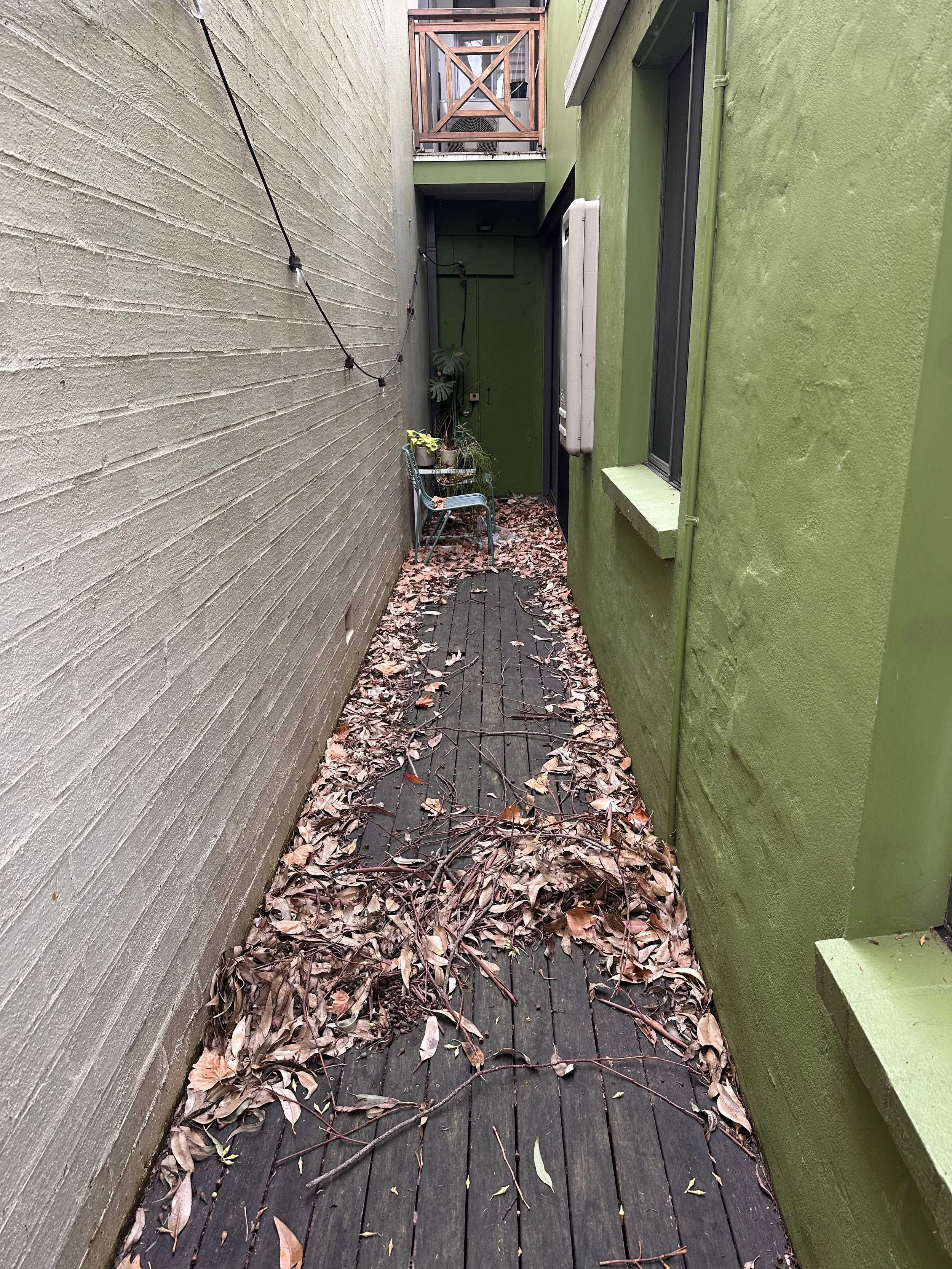An outdoor narrow pathway with a wooden deck and green and beige exterior walls. The path is covered with fallen dried leaves and twigs, and has a small green chair, potted plants, and a string of lights.