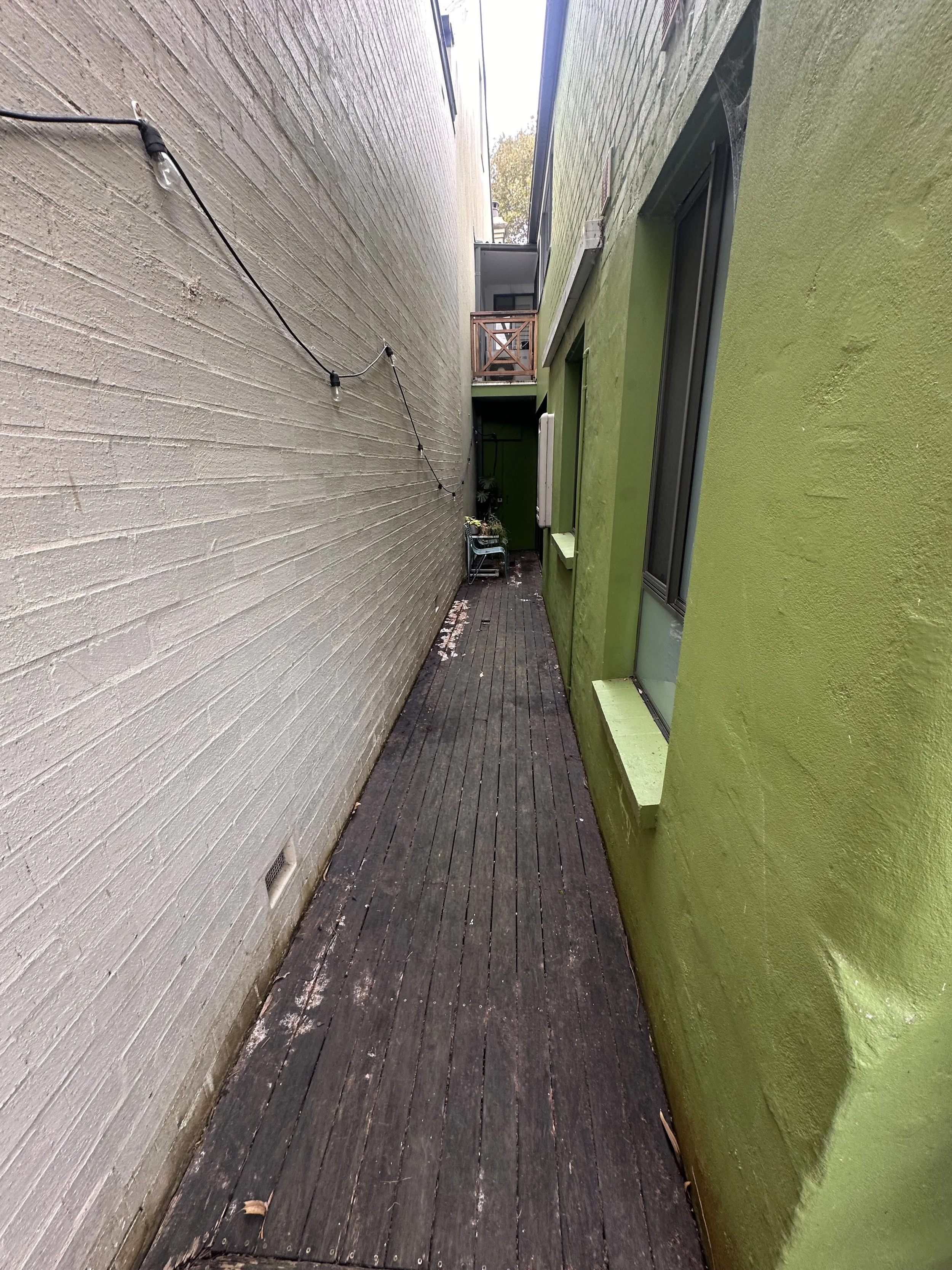 A narrow alleyway with a wooden deck floor, flanked by a beige painted wall on the left and a green painted wall on the right. There is a string of outdoor lights attached to the beige wall, and some potted plants and patio furniture in the distance.