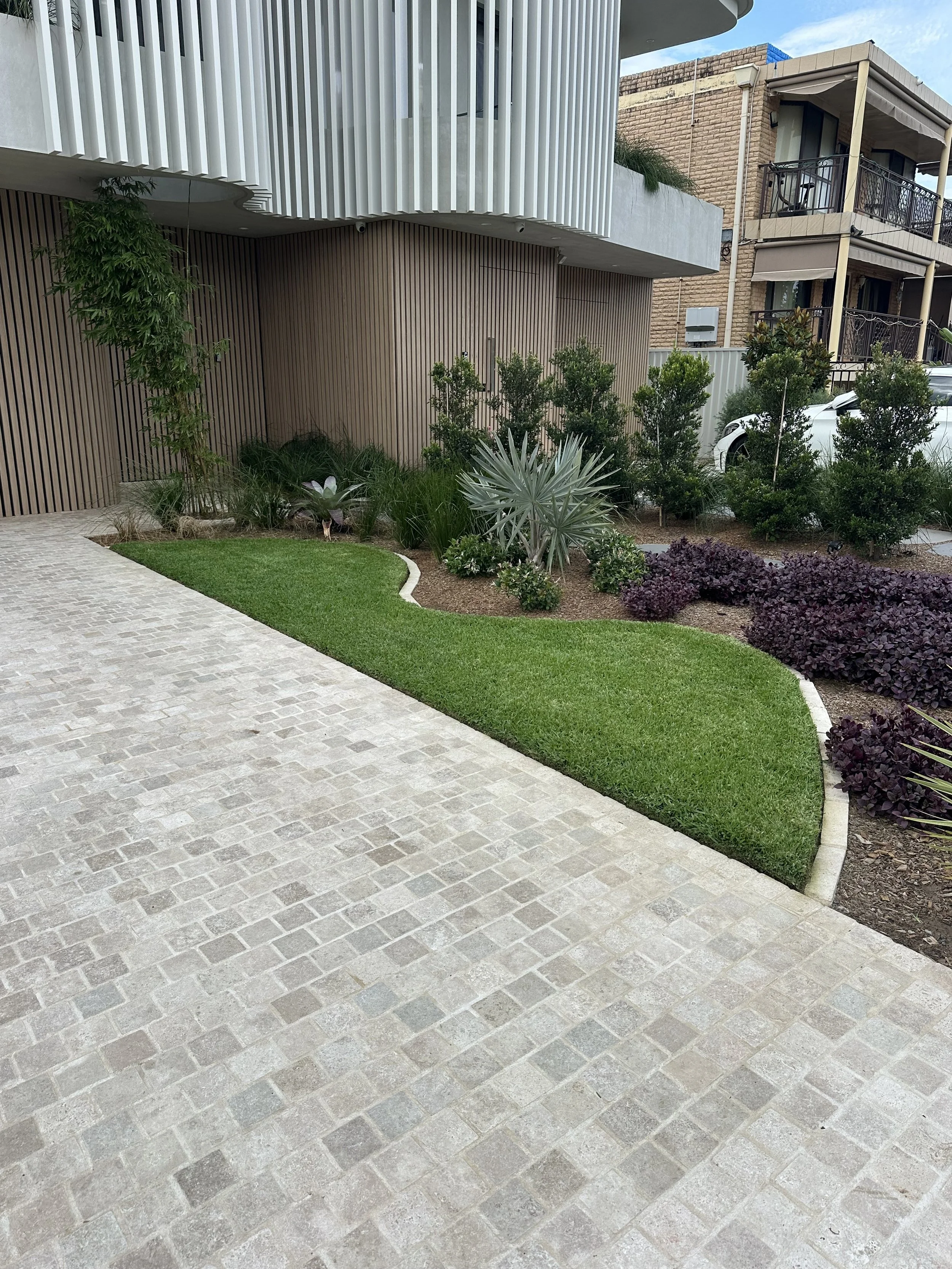 Modern residential building with a landscaped garden, concrete pavers, green grass, various plants, and bushes. A car is parked on the driveway.