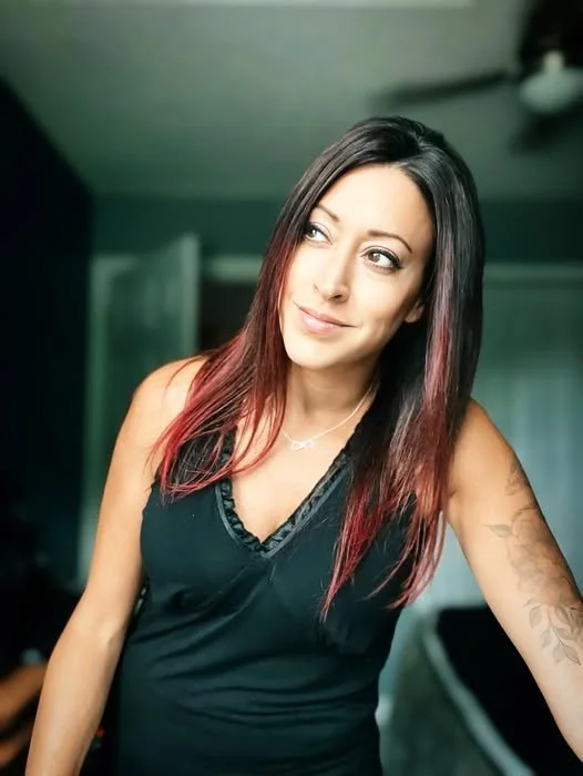 A woman with long, dark hair that has red tips, wearing a black sleeveless top, standing indoors with a relaxed expression and slight smile.
