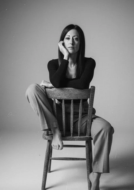 Black and white photo of a woman sitting on a wooden chair facing forward, with her right elbow resting on the top of the chair back and her right hand supporting her face. She is wearing a long-sleeved black top and light-colored loose-fitting pants, with her left foot resting on the seat of the chair and her right foot on the floor.