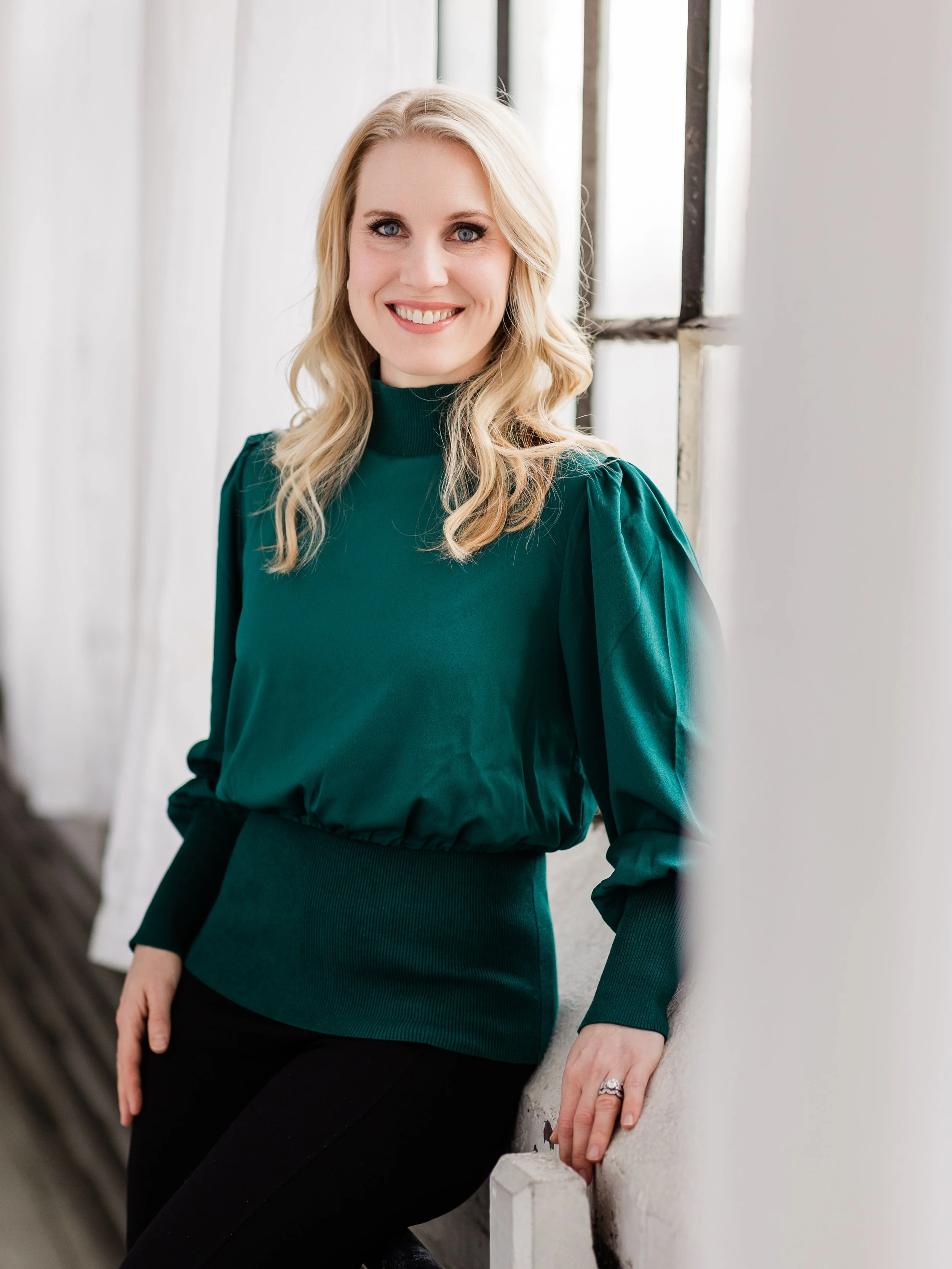 A woman with blonde hair and blue eyes, wearing a green long-sleeve top and black pants, sitting against a white wall near a window.