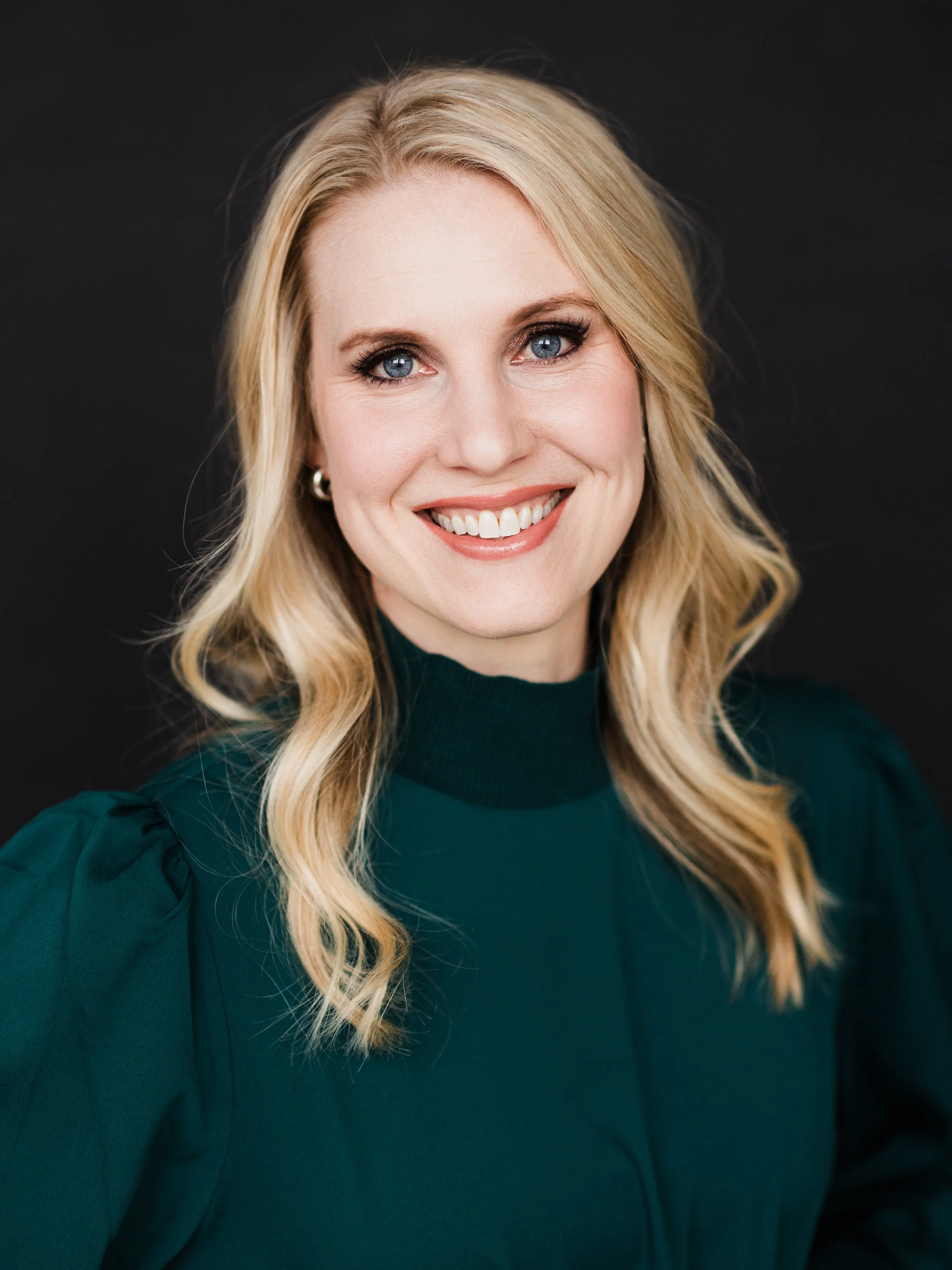 Close-up of a woman with long blonde hair and blue eyes, smiling, wearing a dark green top with puffy shoulders, against a black background.