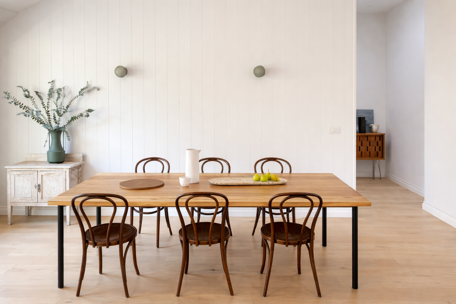 Part of a dining room with a rectangular wooden table and six wooden chairs, a white cabinet with a green vase and leafy plant, and minimal decorative items on the table.