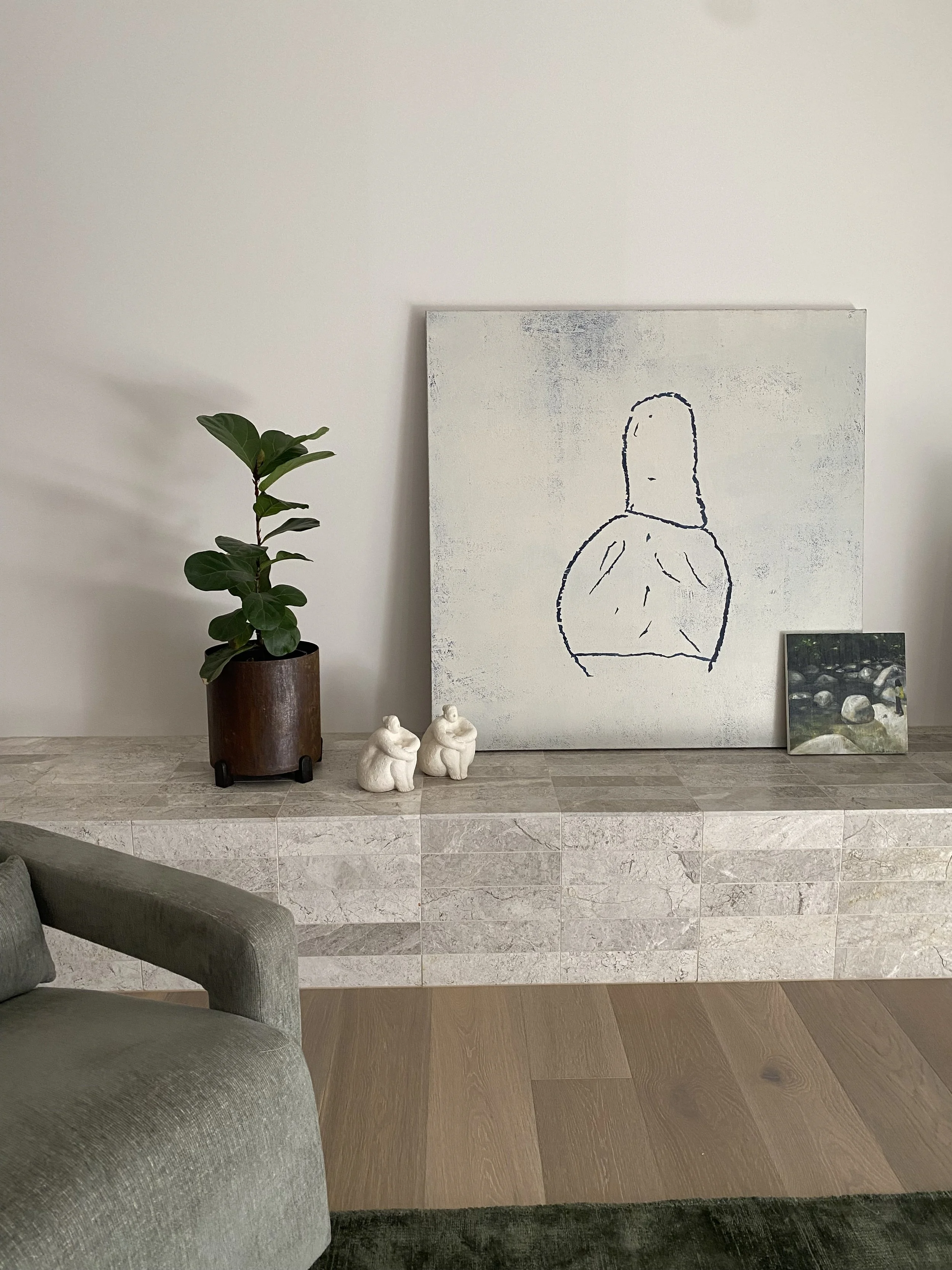Interior of a modern room with a potted plant, abstract line art painting, smaller landscape photo, two ceramic figurines, and a gray armchair in the foreground.