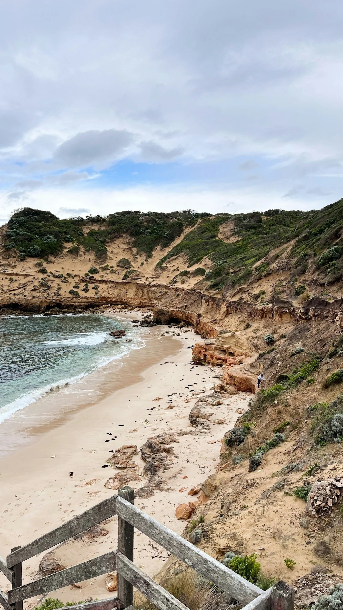 A coastal beach with sandy shores, rocky cliffs, and a person walking along the cliffs, with green shrubbery and rocks.