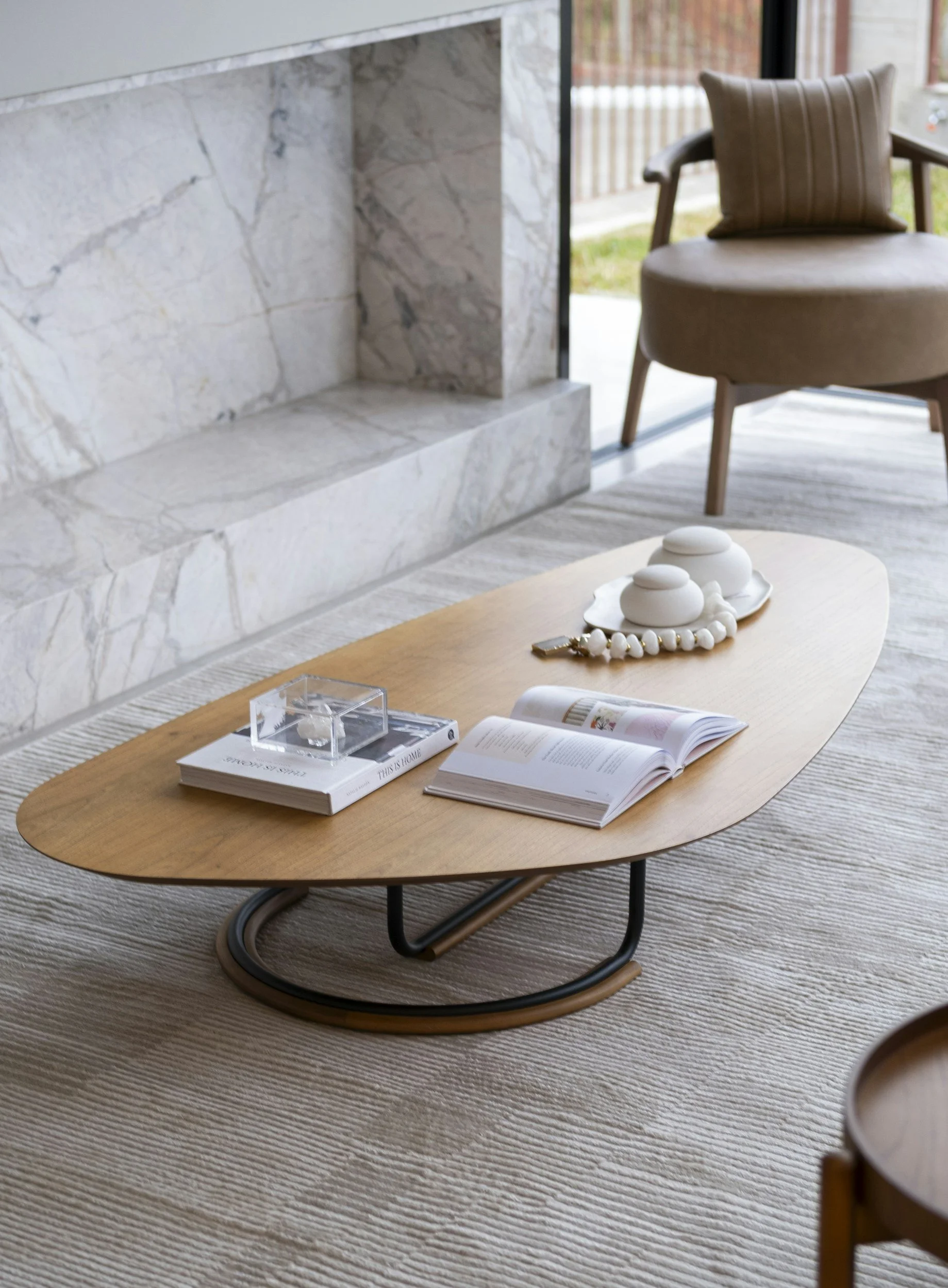 A modern living room with a wooden coffee table on a light-colored rug, a beige armchair with a pillow, a marble fireplace, and a sliding glass door leading outside.