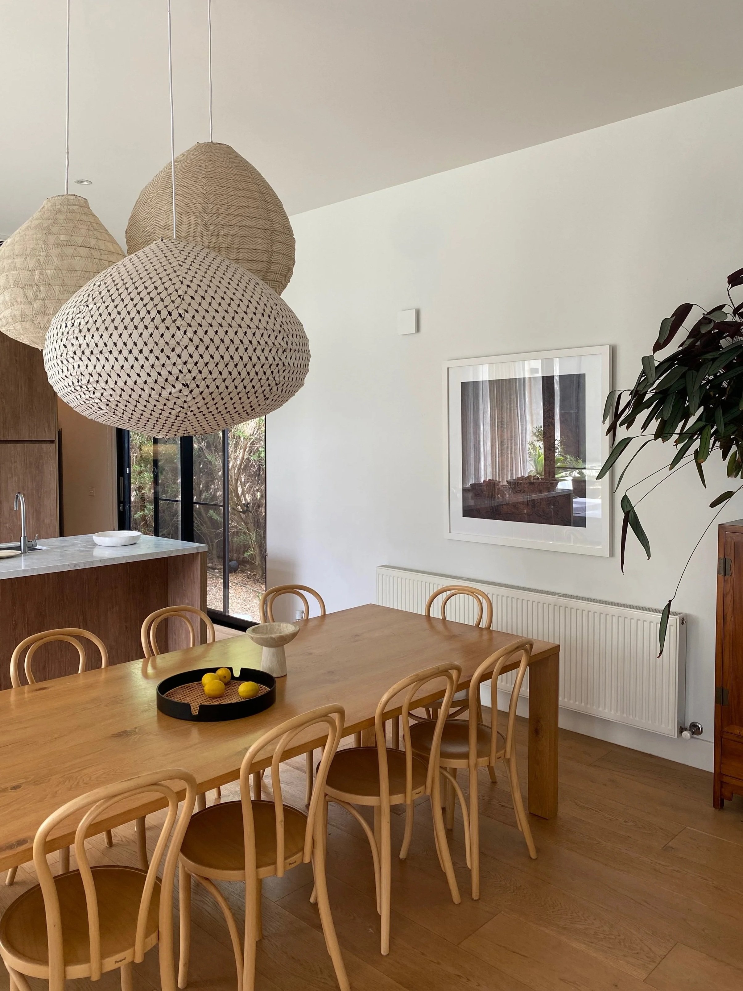 A dining room with a wooden table surrounded by matching wooden chairs, a black tray with yellow lemons, hanging woven pendant lights, a framed artwork on the white wall, and a large plant on the right side.