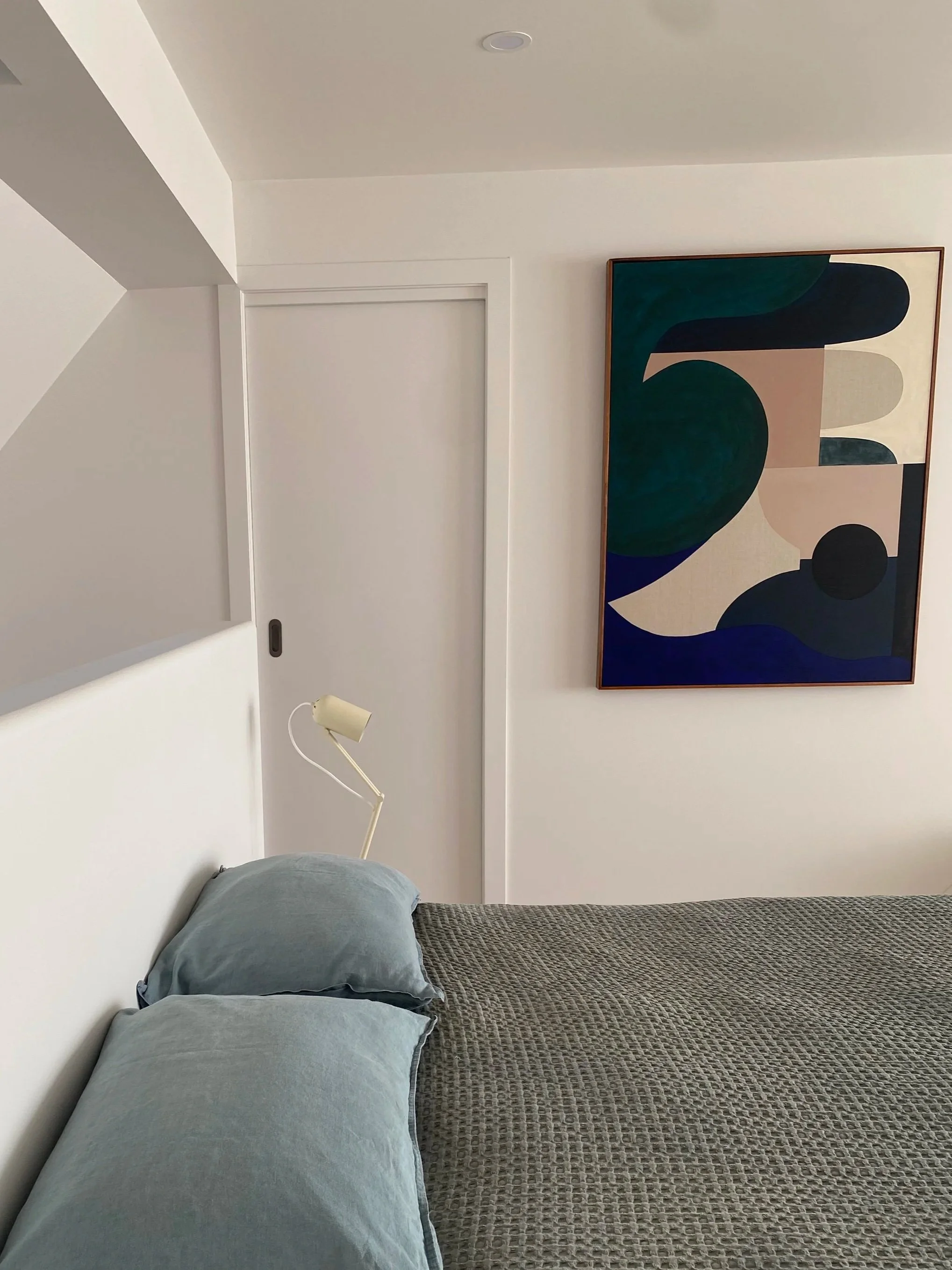 A bedroom with a bed that has a beige textured blanket and light blue pillows. There is a wall-mounted lamp above the bed and abstract art on the wall featuring green, black, beige, and blue shapes. The room has white walls, a sliding door, and a small closet.