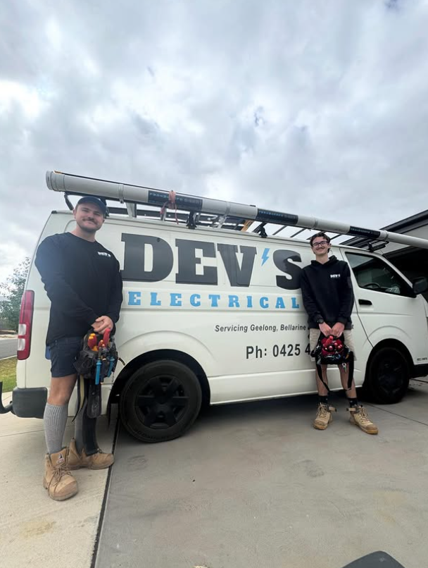 Two electricians in black hoodies standing in front of a white service van with a ladder on top, logo reading 'DEV'S ELECTRICAL', parked on a concrete driveway with cloudy skies overhead.