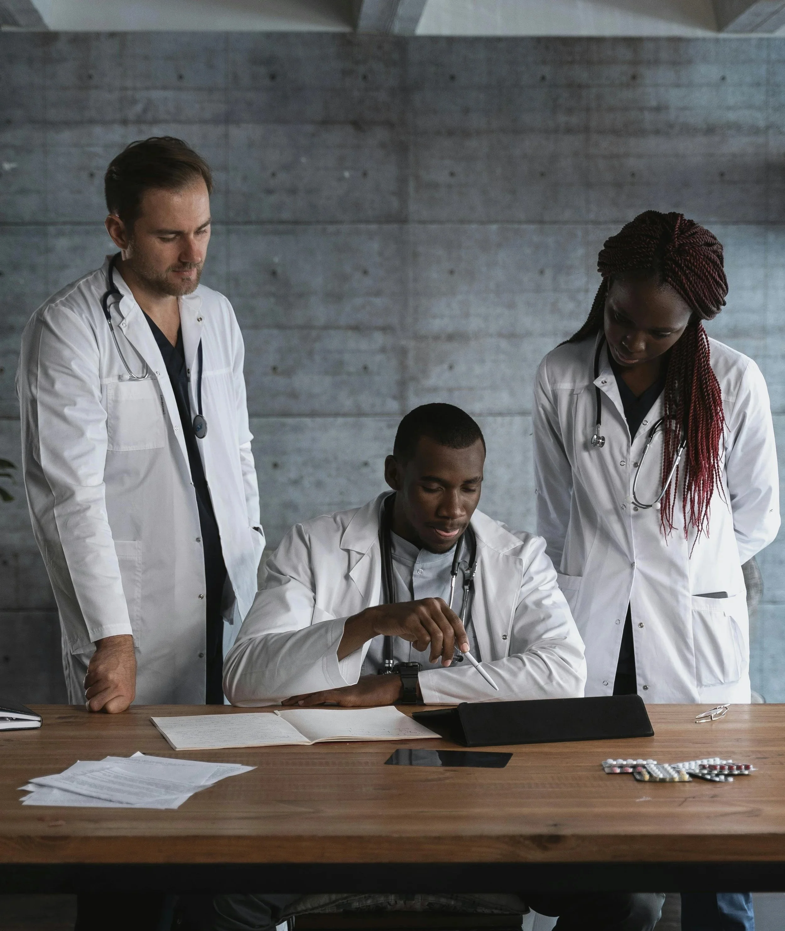Three healthcare professionals, including two women and one man, in white coats gathered around a table looking at documents and a tablet, with a concrete wall in the background.