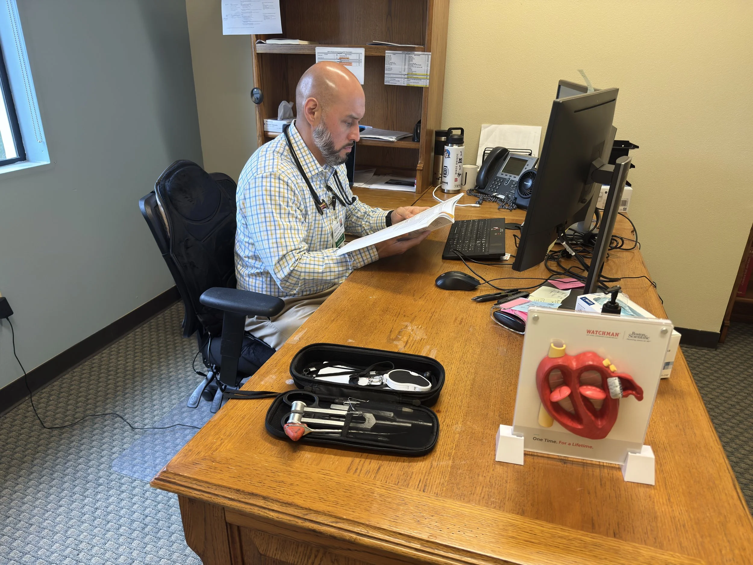 A doctor sitting at a wooden desk in an office, reading a book. The desk has a computer, phone, and medical tools including an otoscope and a reflex hammer. There is a heart model displayed on a stand on the desk.