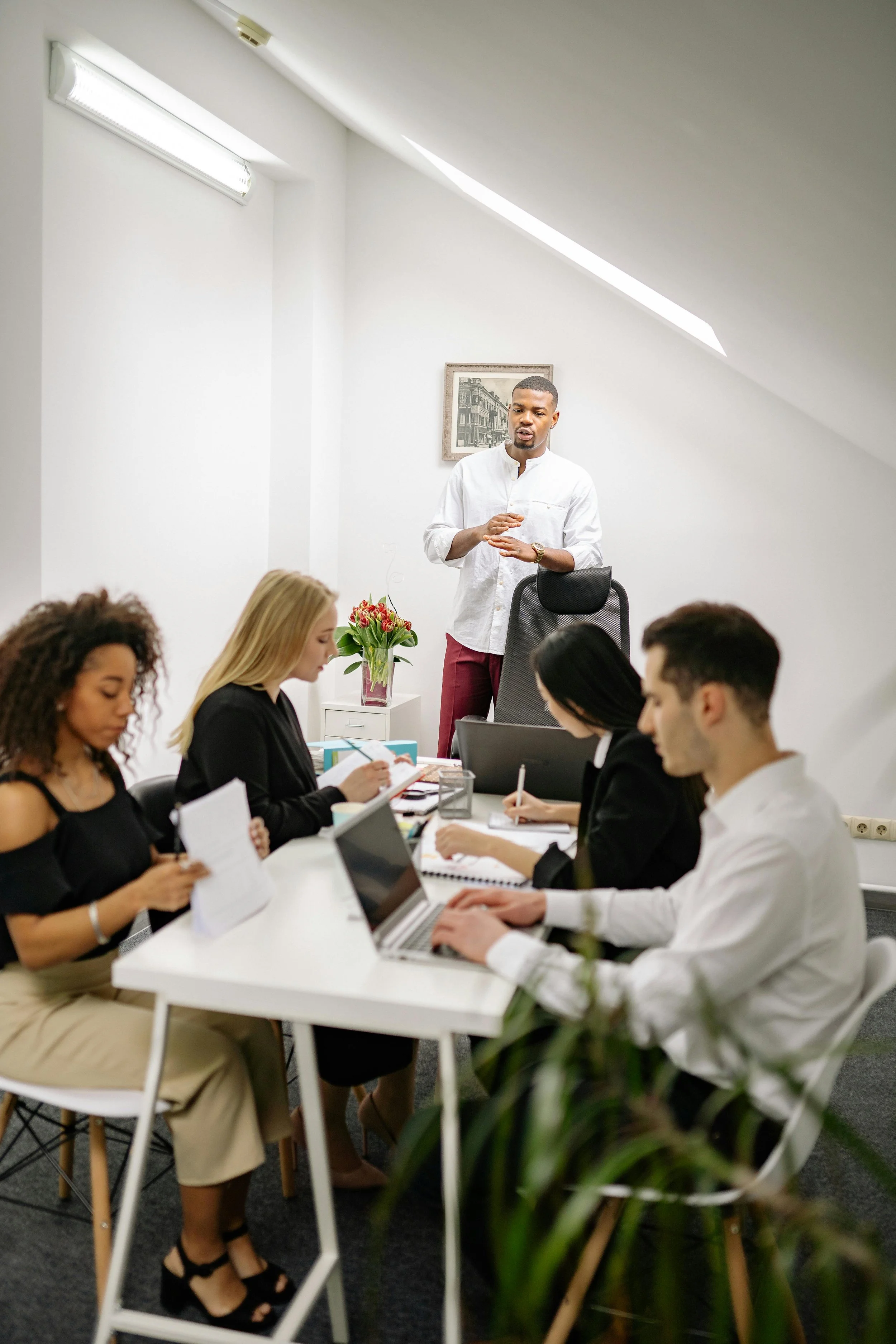 A man in a white shirt standing in front of a group of four people seated at a white table, engaged in a meeting in a bright office with white walls and a skylight.