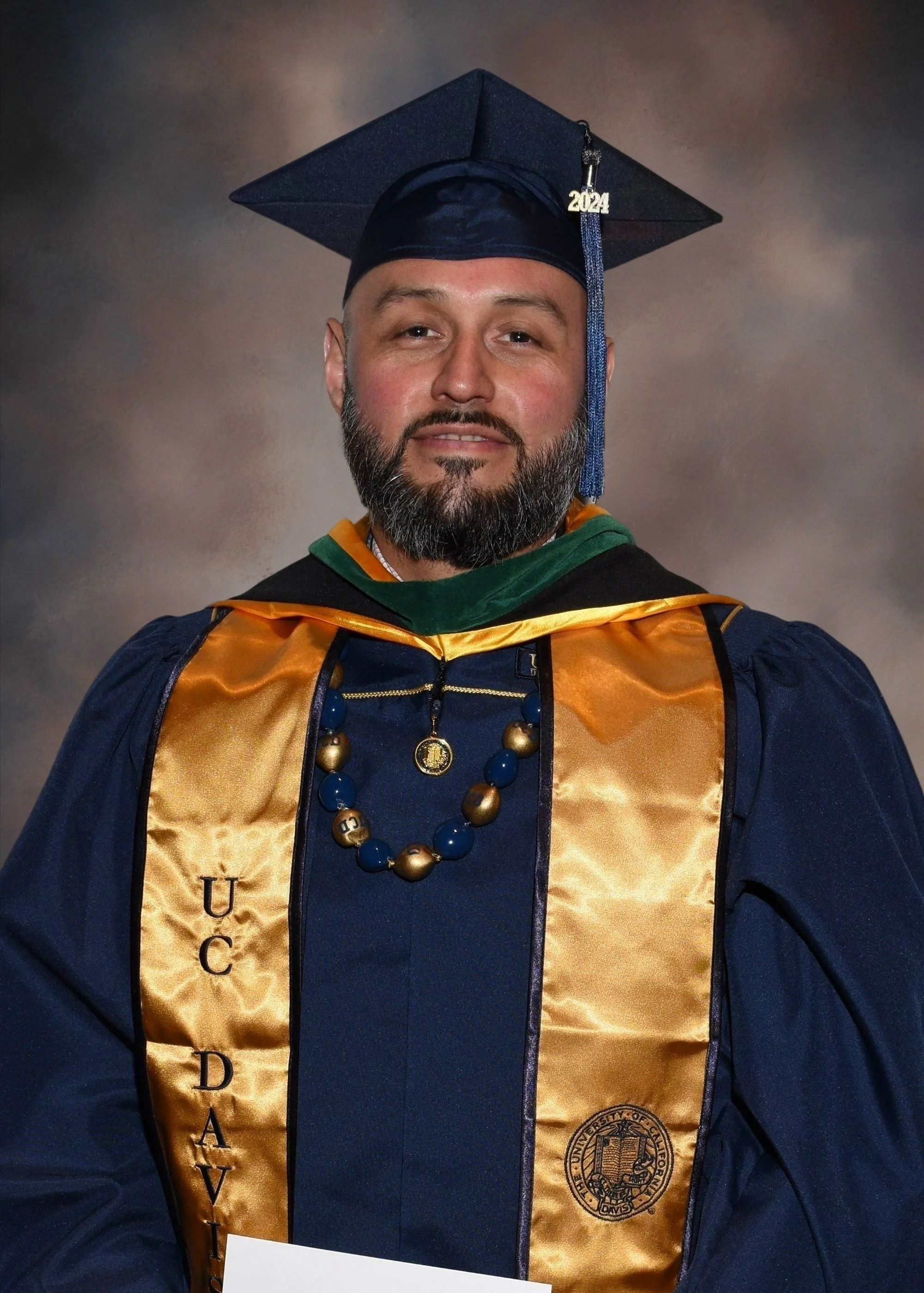 A man in a graduation cap and gown, wearing a gold sash with 'UC DAVIS' written on it, a blue necklace, and a medallion, posed for a graduation portrait.