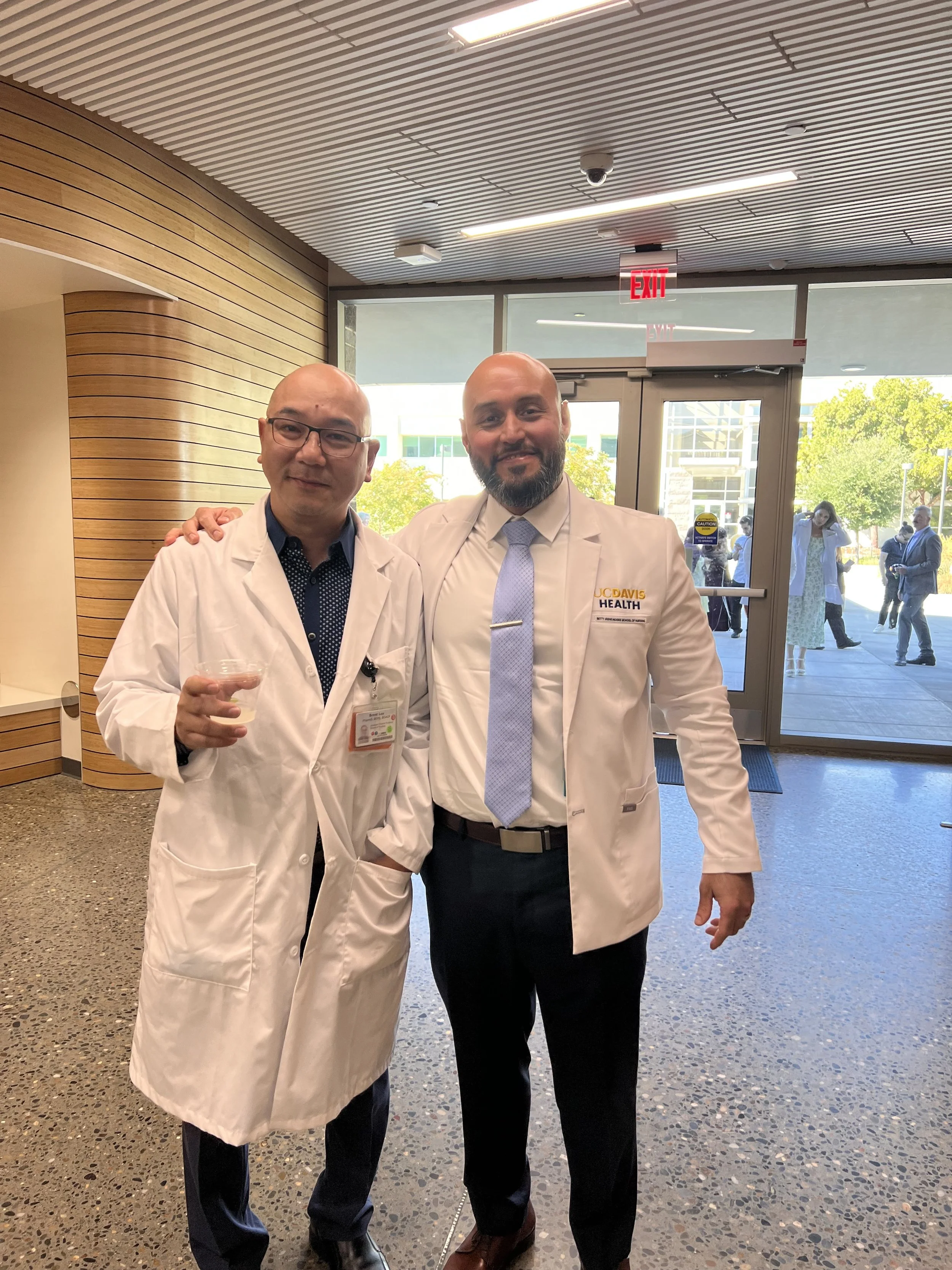 Two men in white medical coats standing inside the hospital near the entrance. One is holding a drink, and both are smiling at the camera.
