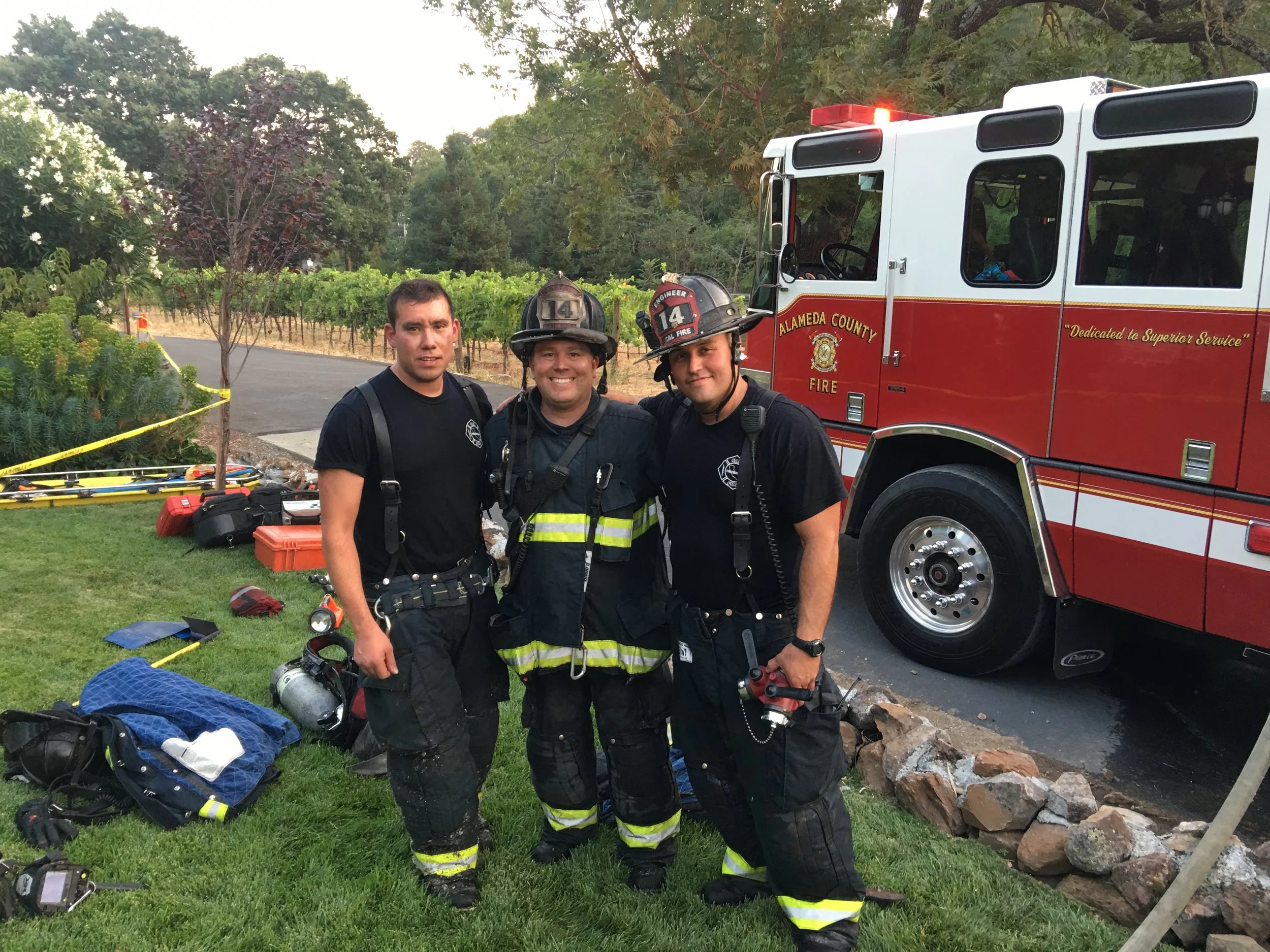 Three firefighters standing in front of a red Alameda County Fire truck, smiling, wearing black uniforms and helmets. Scene includes equipment on the grass and a yellow caution tape, with trees and a vineyard in the background.
